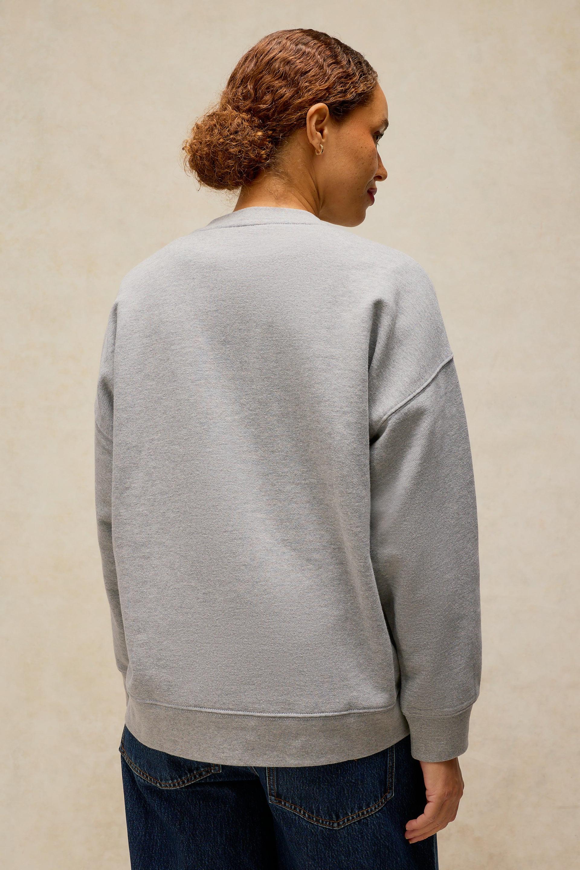 A woman shown from the back wearing a light gray crewneck sweatshirt and dark blue jeans, standing against a textured beige wall.