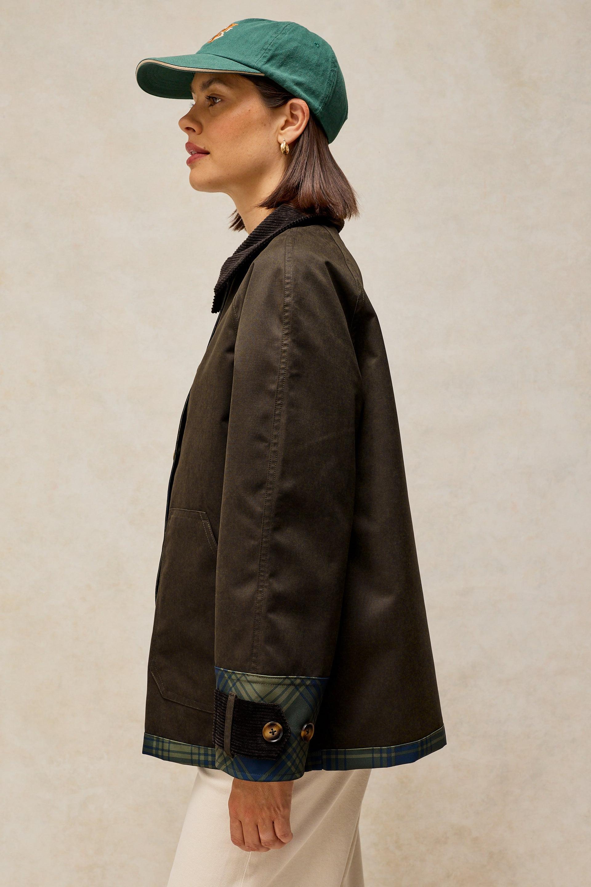 A woman shown in profile wearing a dark brown waxed canvas utility jacket with green plaid cuff lining and a green baseball cap, set against a textured beige background.