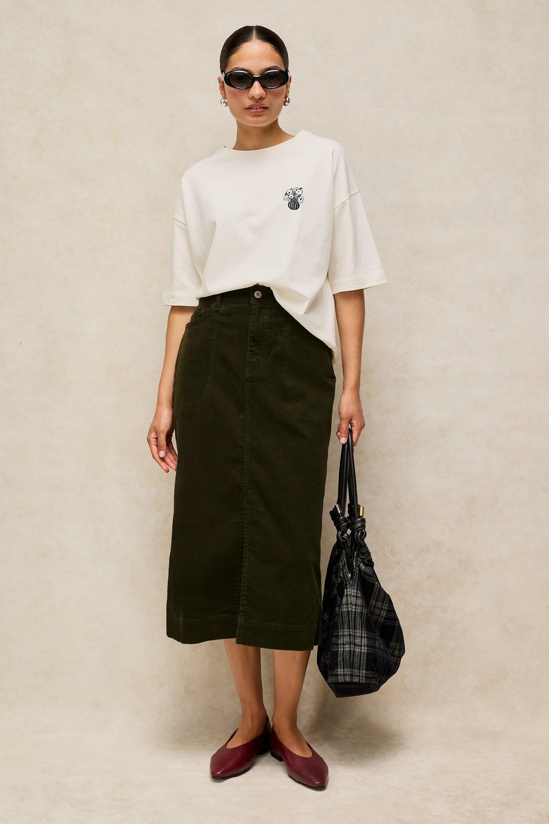 A woman wearing a green cord midi skirt, a white t-shirt, and holding a plaid handbag against a neutral background.