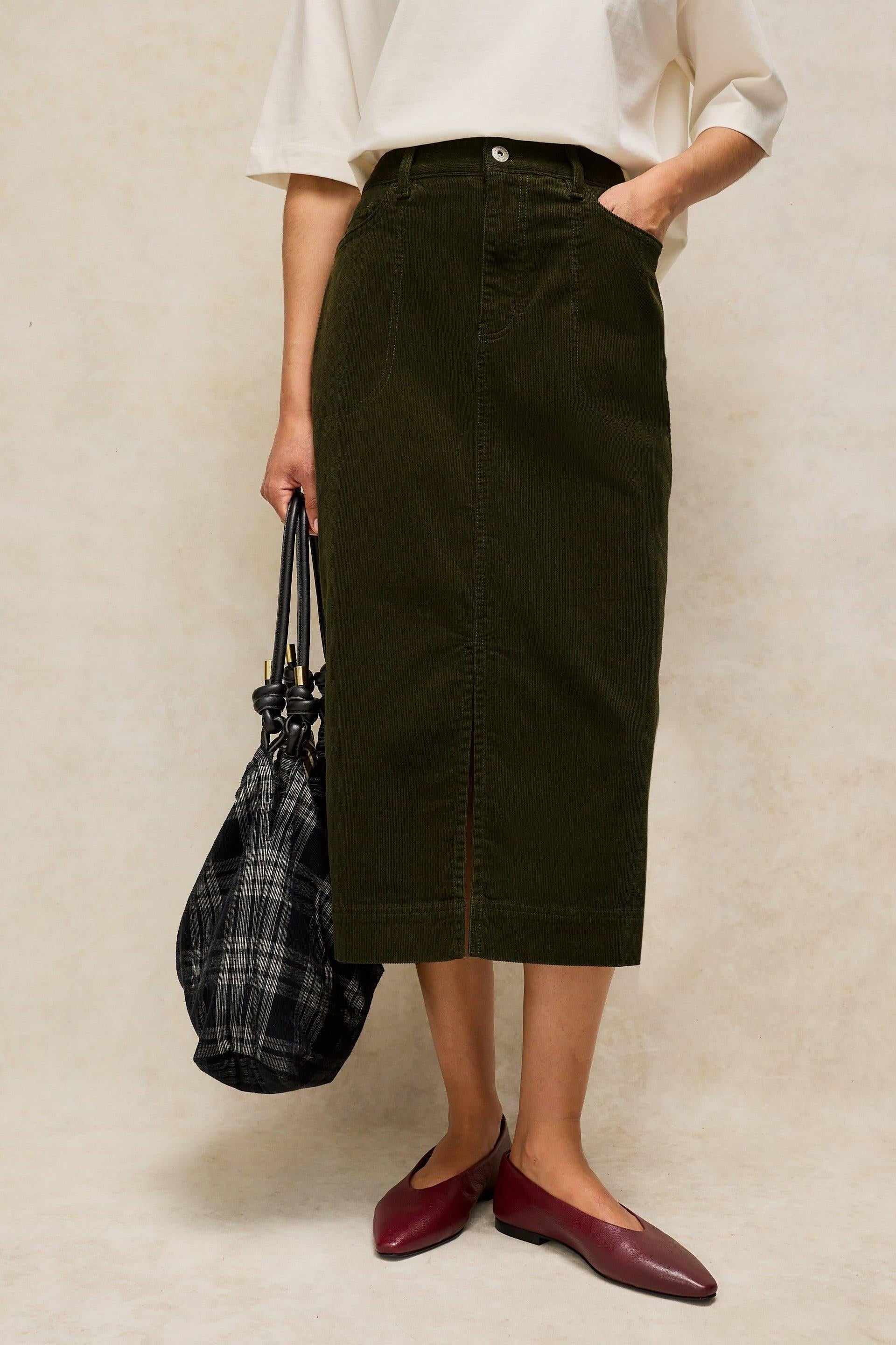 A person wearing a dark green corduroy midi skirt with a white t-shirt and maroon flats.