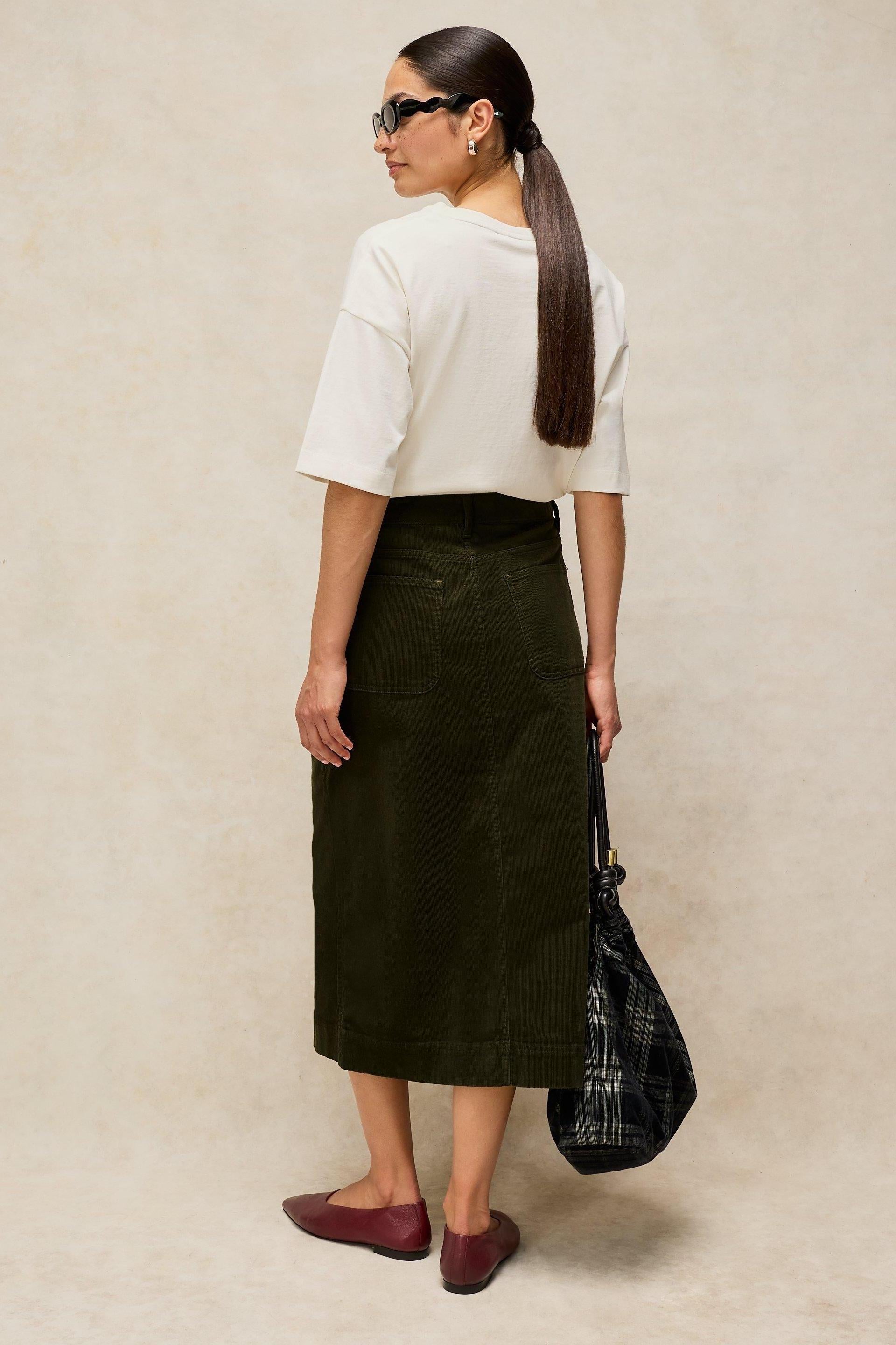 Woman seen from behind wearing a green corduroy midi skirt and a white t-shirt against a neutral background.