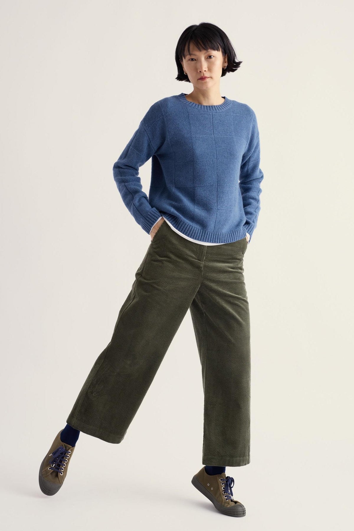 A woman wearing olive green cropped corduroy trousers and a blue textured knit sweater against a plain background.