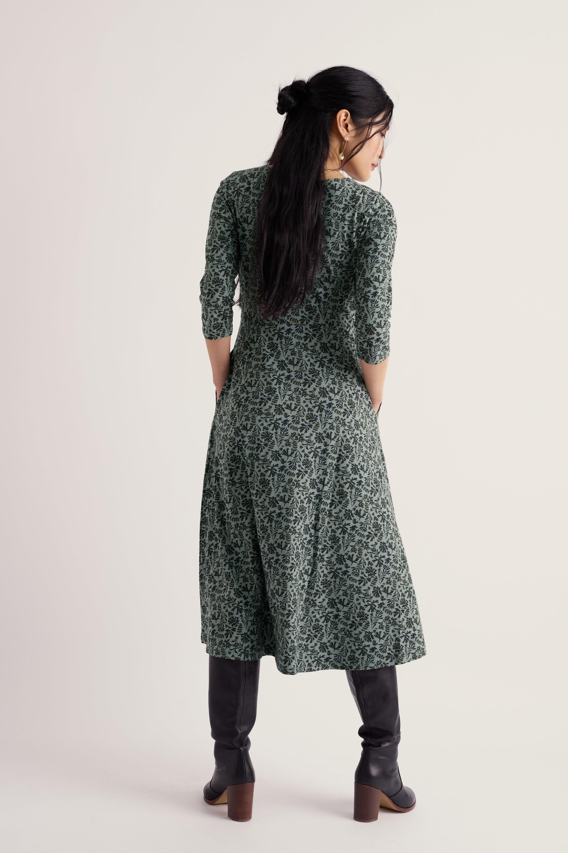A woman viewed from the back wearing a dark green long-sleeve midi dress with a dense small floral print, paired with black knee-high boots, against a plain light beige studio background.