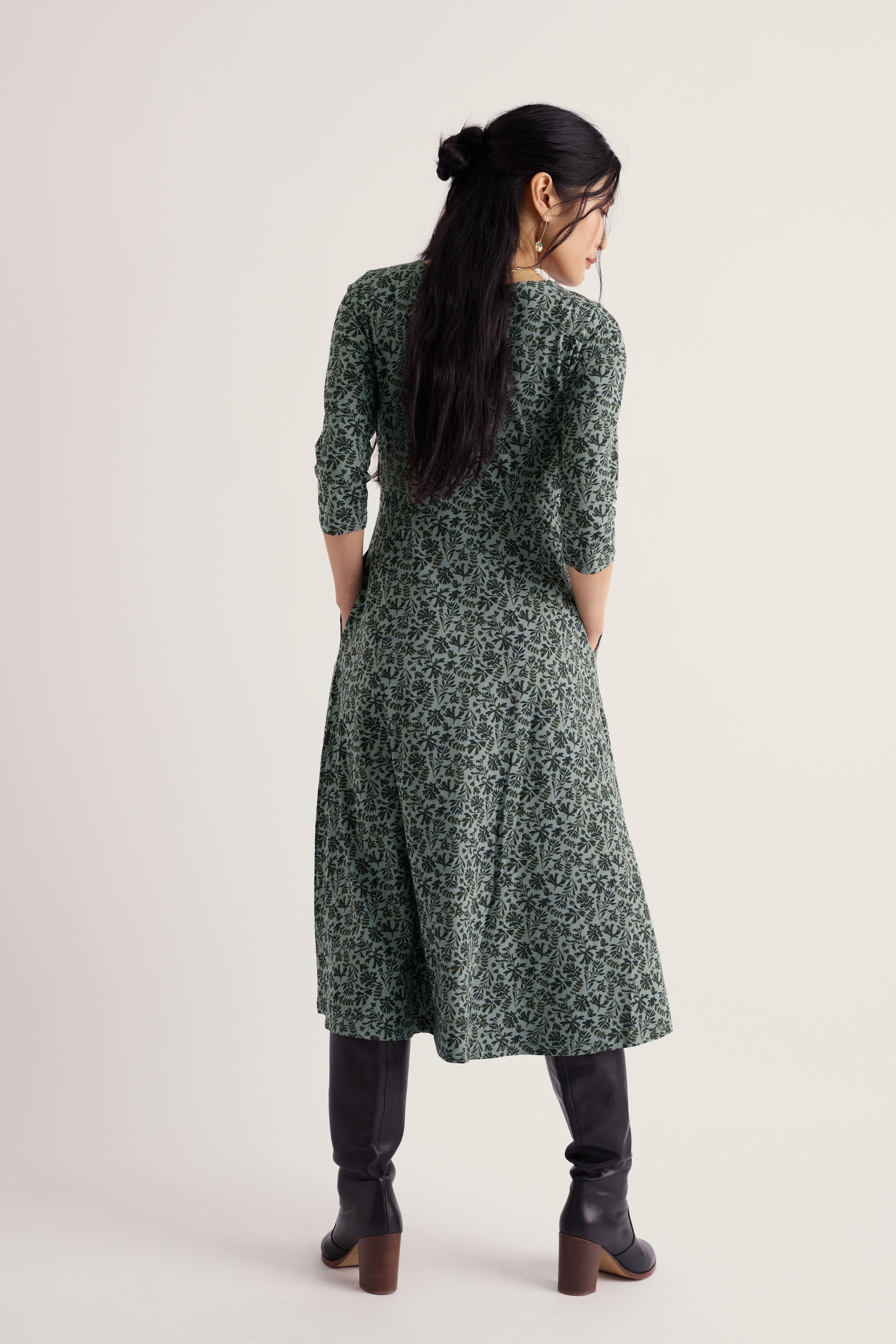 A woman viewed from the back wearing a dark green long-sleeve midi dress with a dense small floral print, paired with black knee-high boots, against a plain light beige studio background.