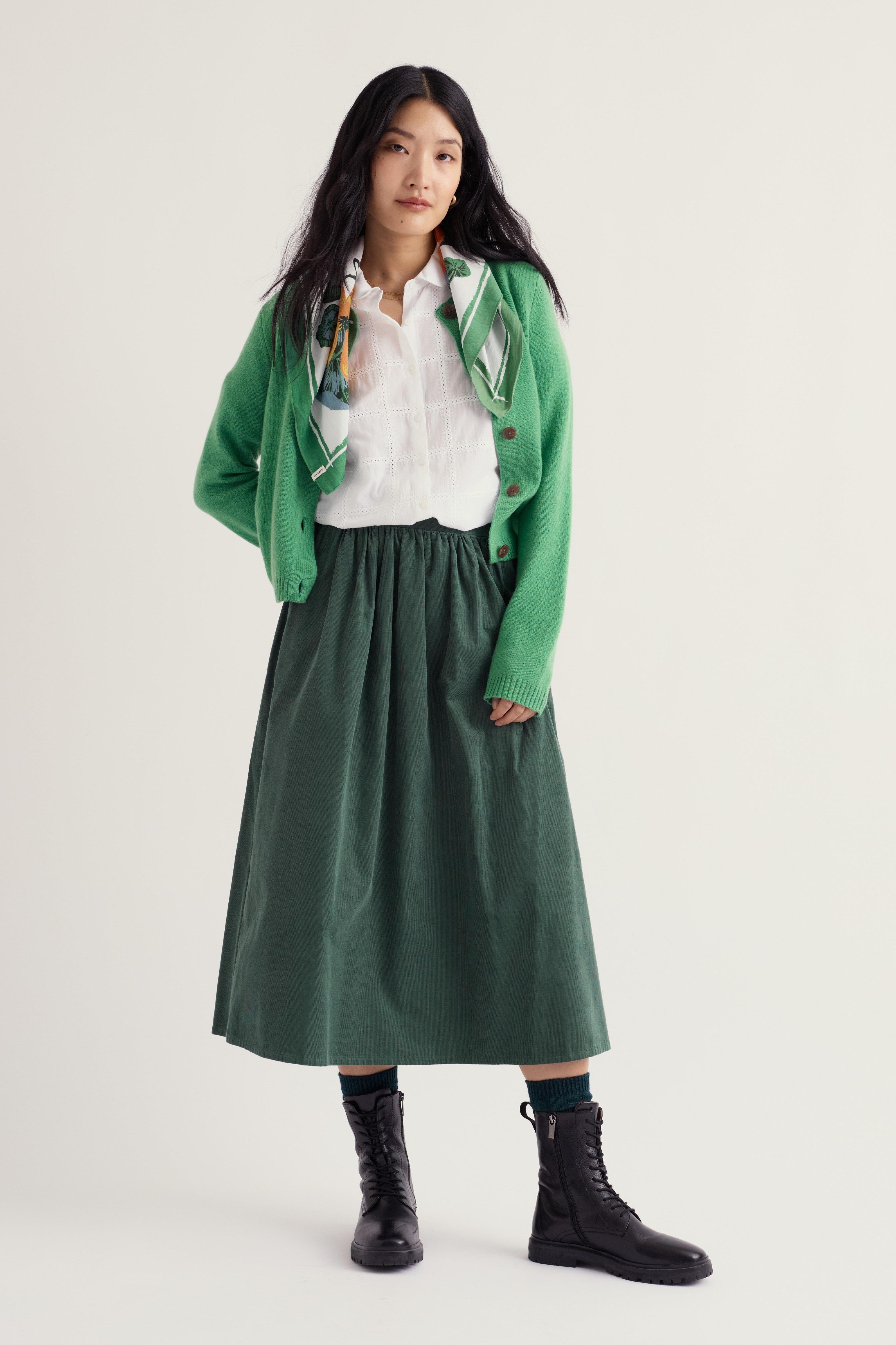 A model stands against a light off-white studio background wearing a green knit cardigan over a white collared shirt, a dark green pleated midi skirt, dark socks, and black lace-up ankle boots.