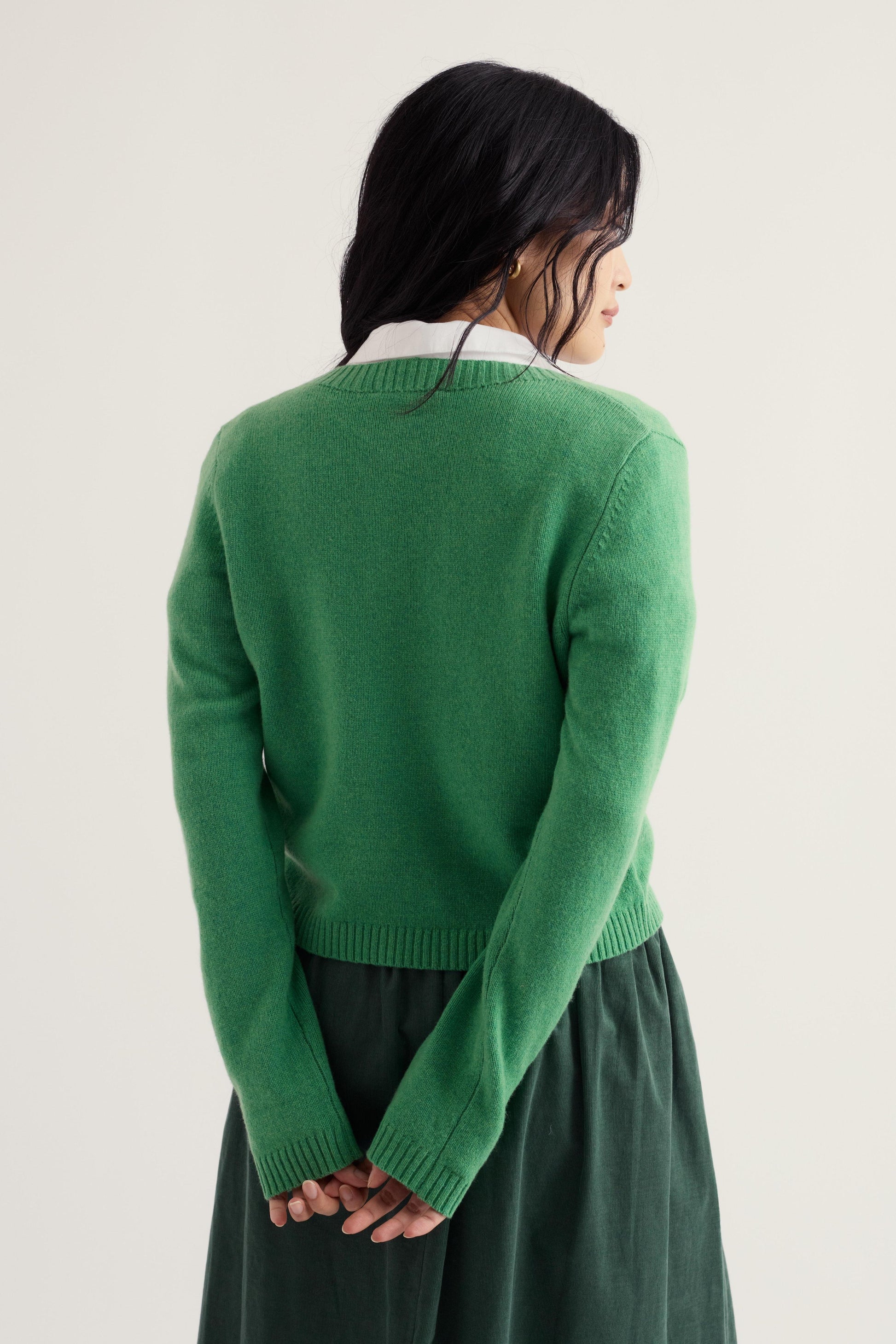 A model shown from the back wearing a bright green knit cardigan over a white collared shirt and a dark green skirt, set against a plain off-white studio background.