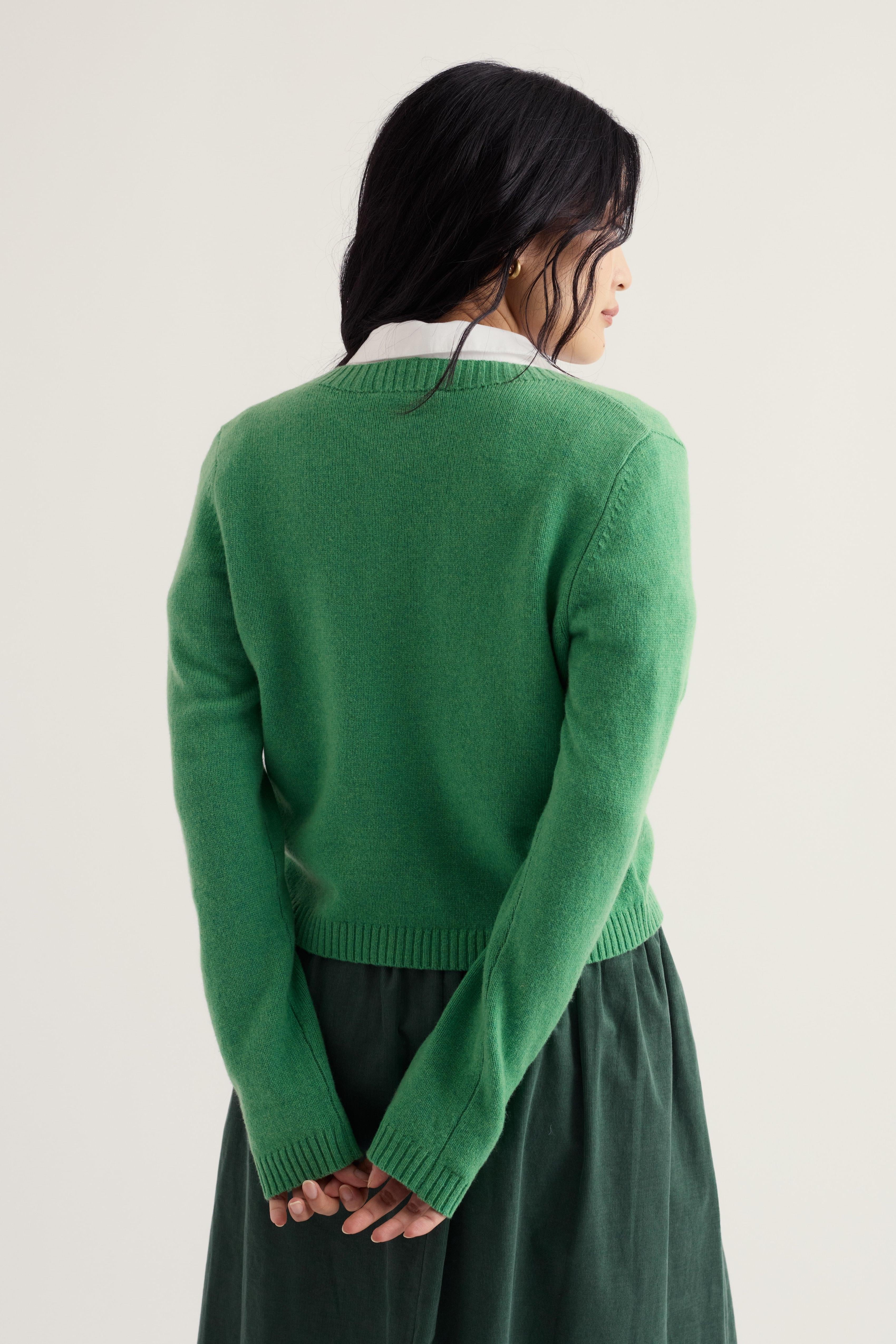A model shown from the back wearing a bright green knit cardigan over a white collared shirt and a dark green skirt, set against a plain off-white studio background.