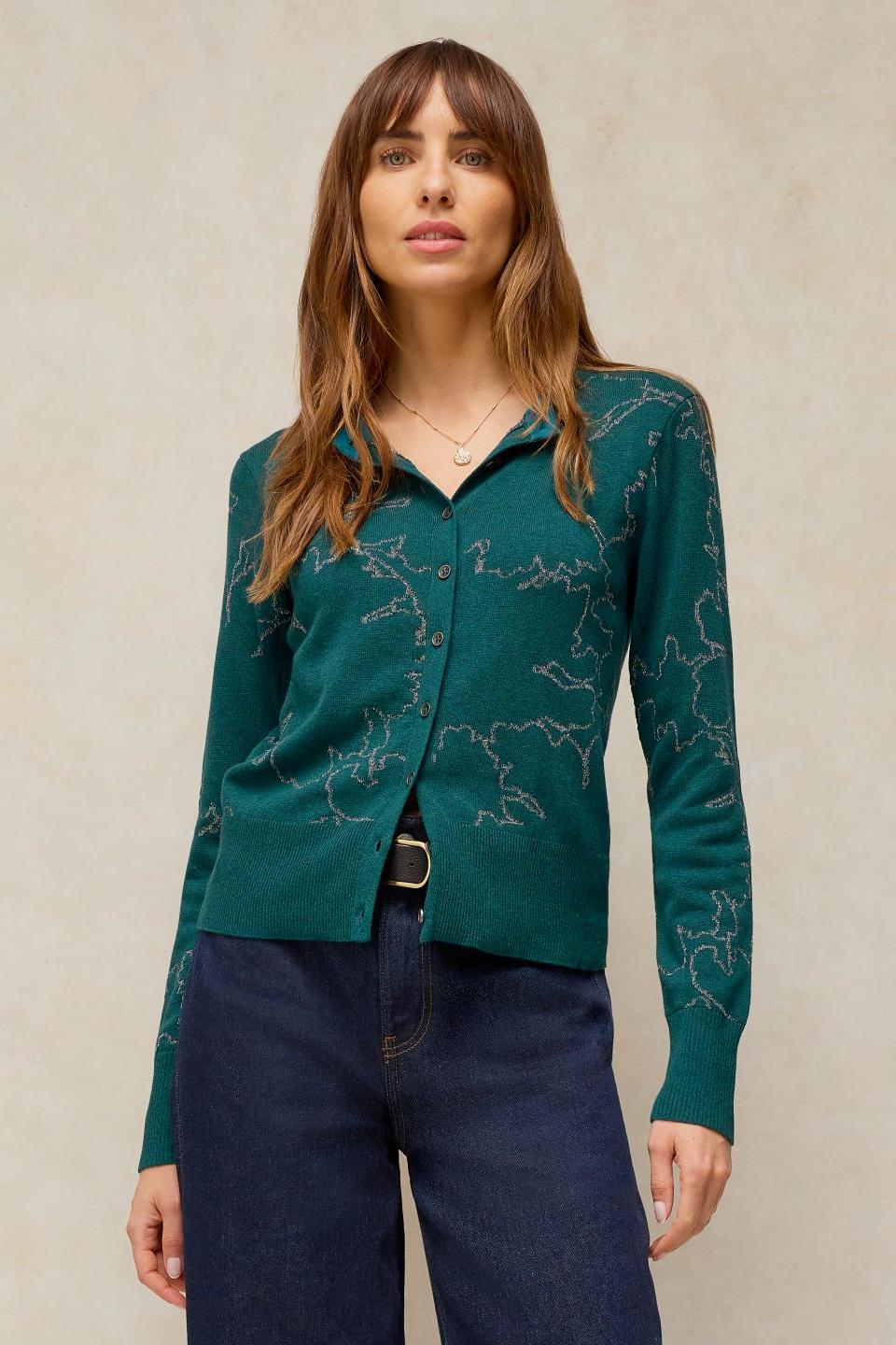 A woman wearing a dark green, metallic-patterned button-down knit cardigan paired with dark blue jeans.