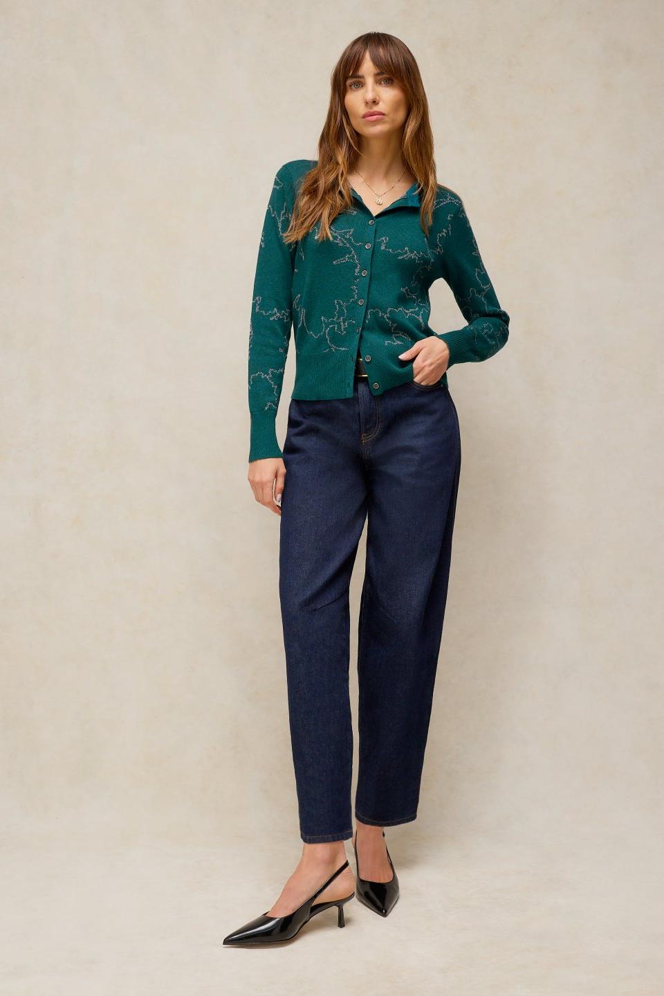 A woman modeling a green metallic knit cardigan paired with dark denim jeans against a neutral studio background.