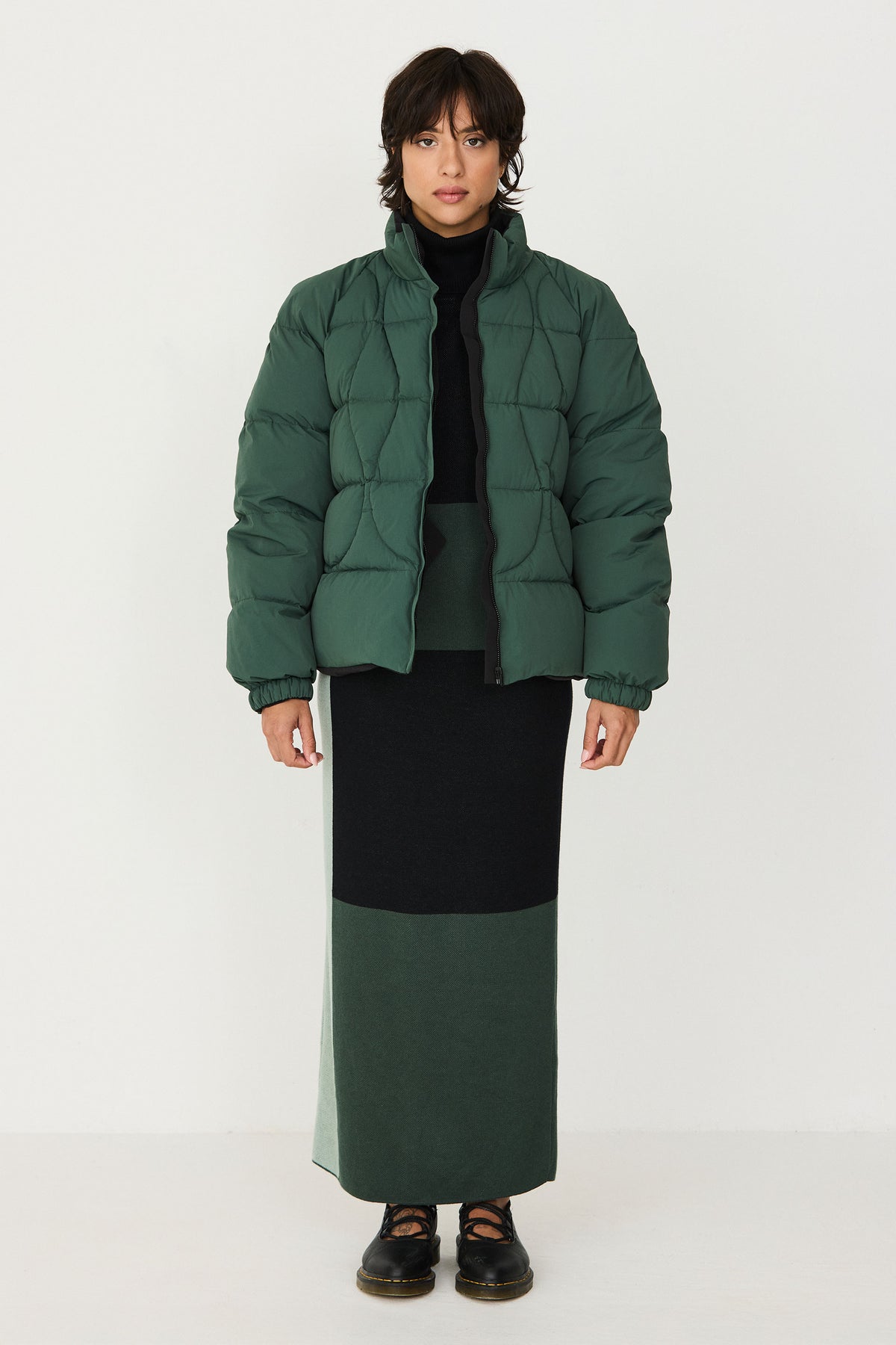 Woman wearing a dark green puffer jacket over a black and green colour-blocked cotton knit ankle length skirt.