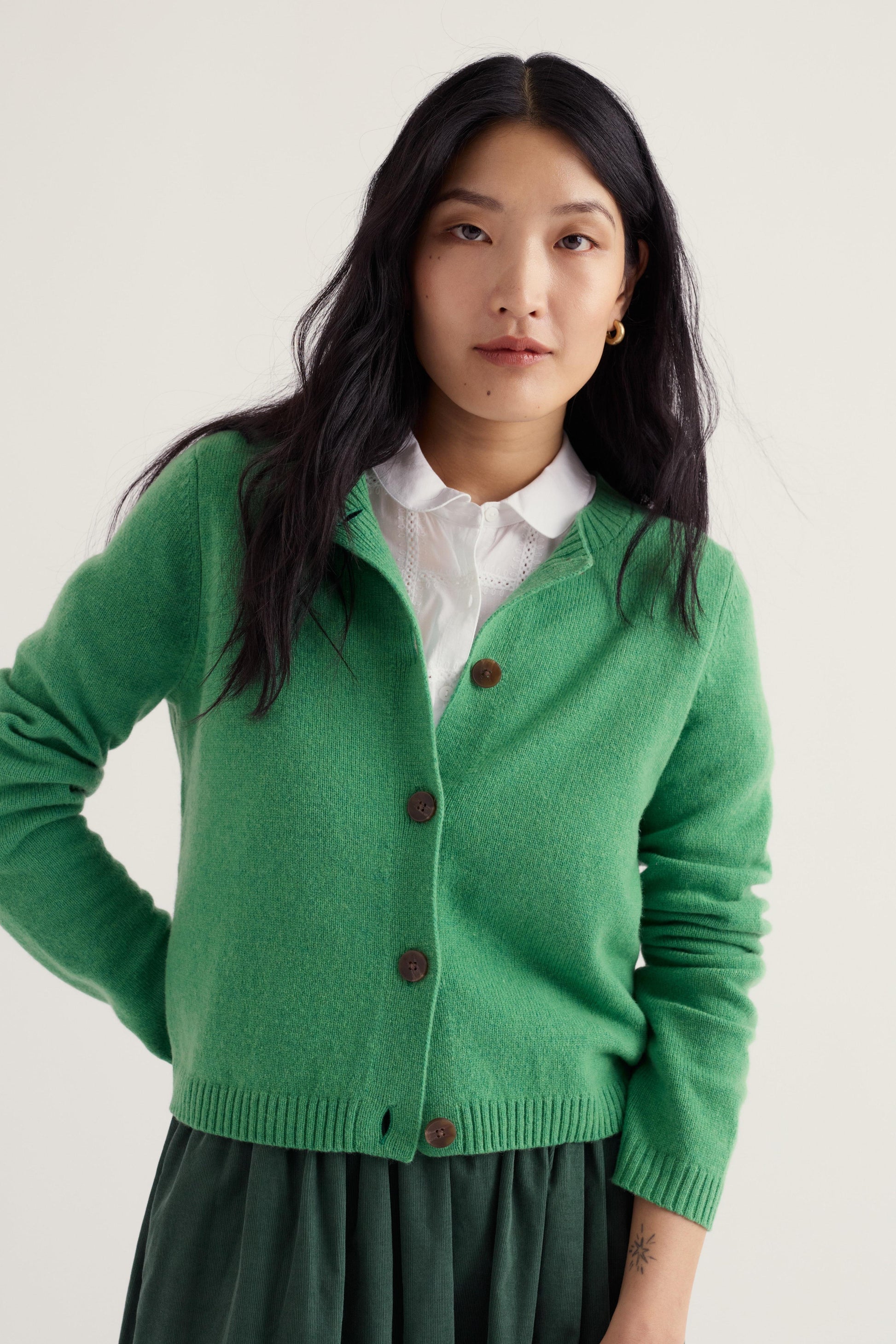 A woman wearing a bright green wool cardigan over a white collared shirt and a dark green skirt, photographed against a plain light beige studio background.