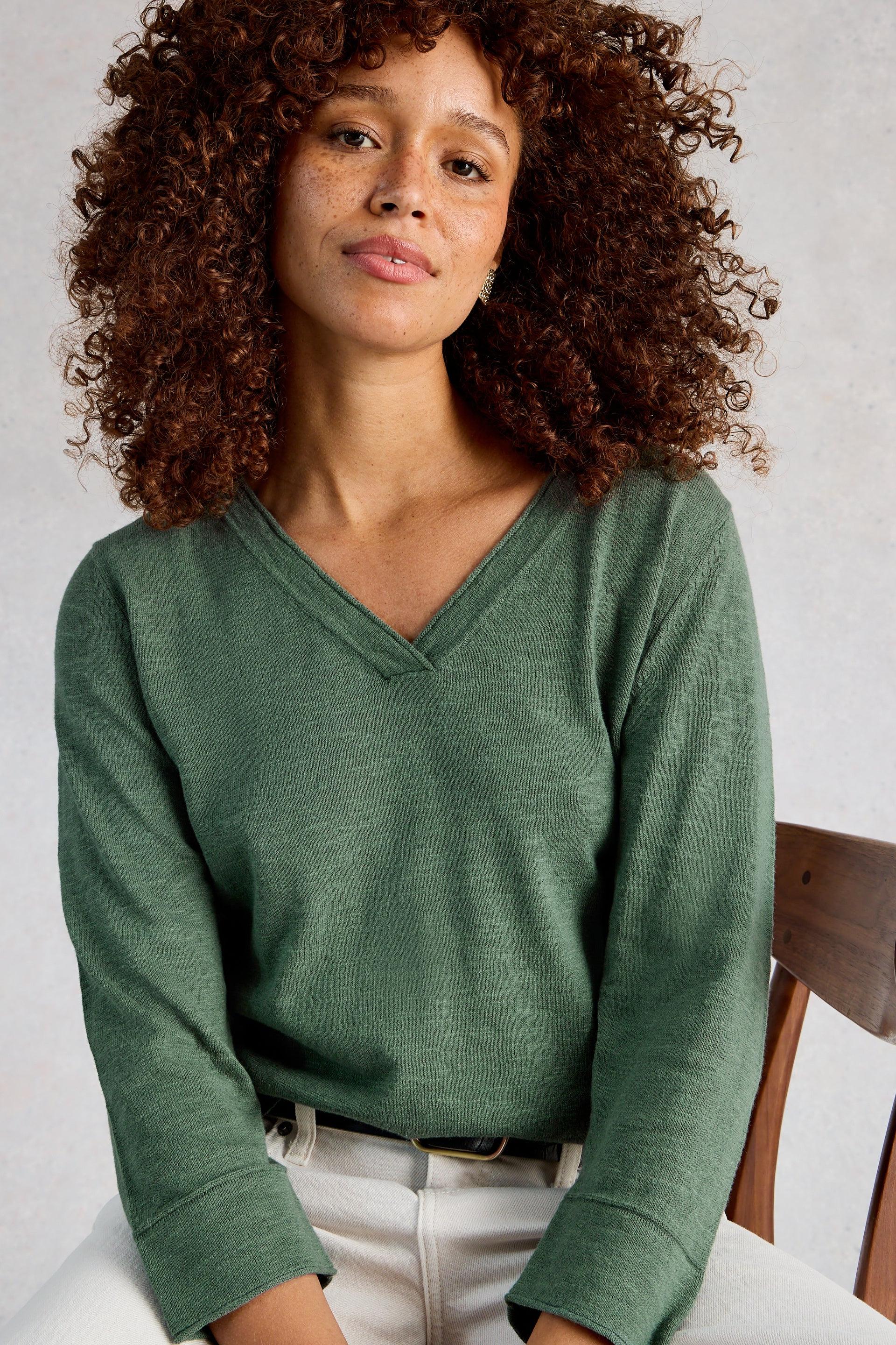 Woman with curly hair wearing a green v-neck organic cotton sweater seated on a wooden chair.