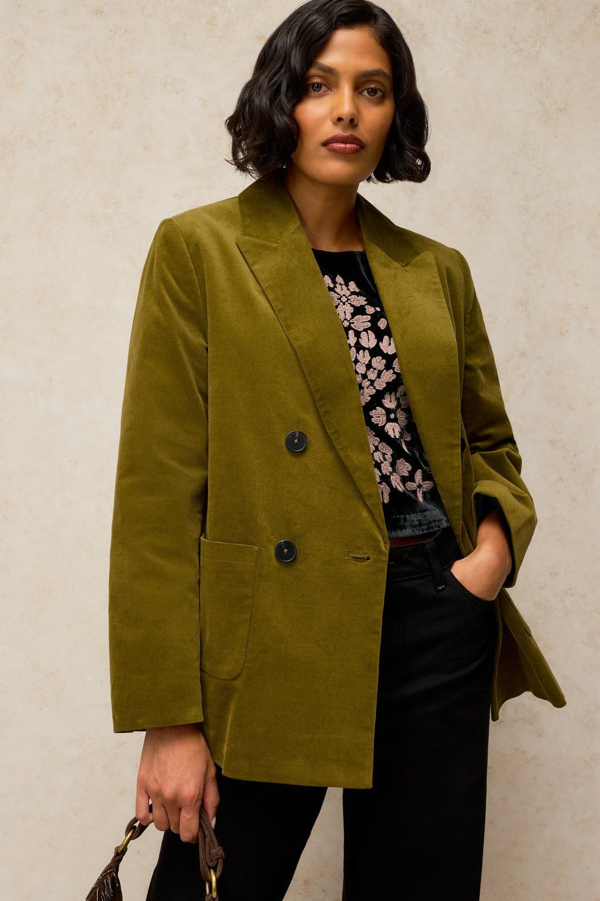 A woman wearing a double-breasted green velvet blazer over a floral embroidered top and black trousers.