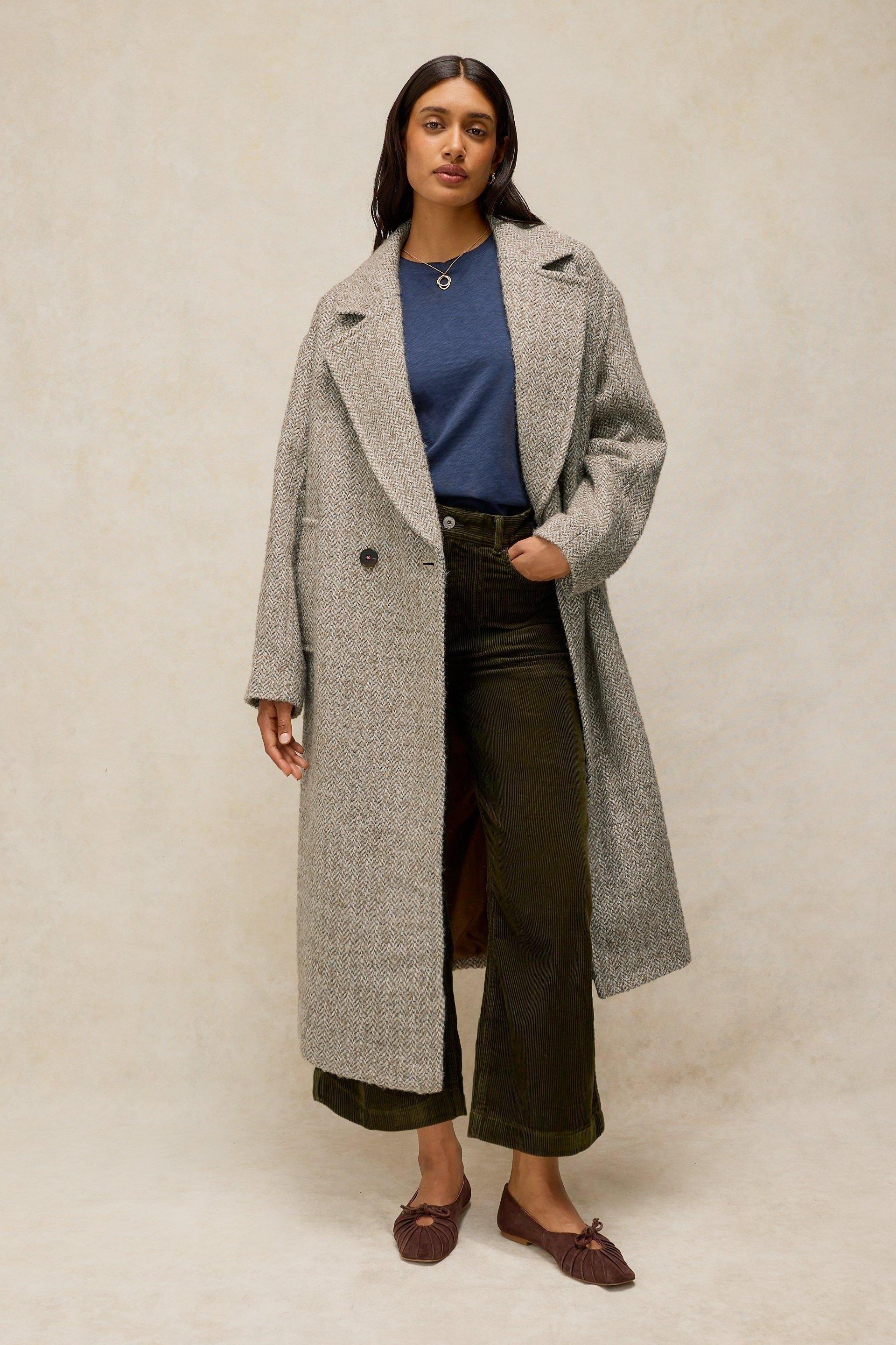 A woman modeling a long herringbone women's wool coat over a blue top and olive green corduroy trousers.