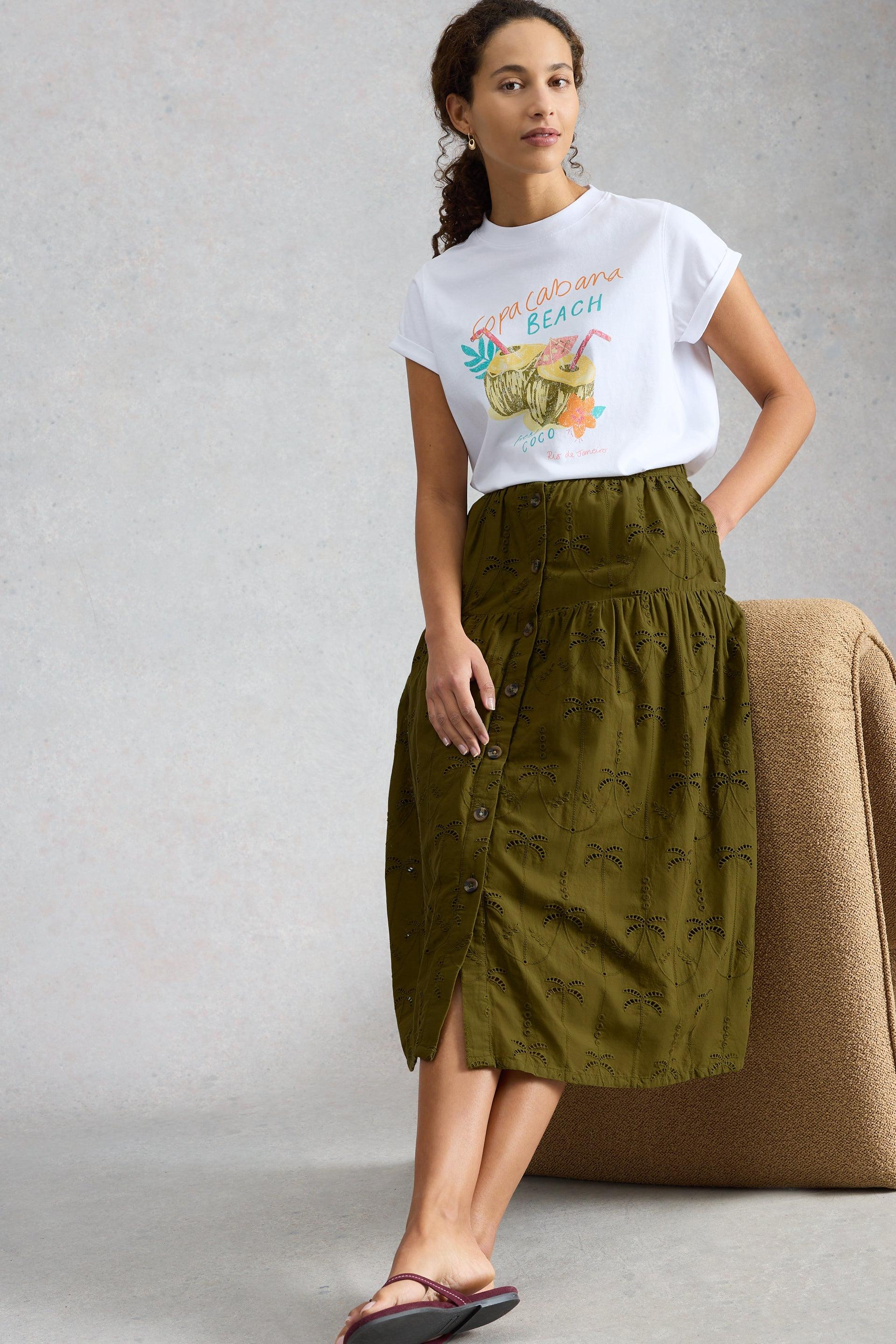 Woman wearing a button-front olive green broderie skirt and Copacabana graphic t-shirt leaning against a chair.