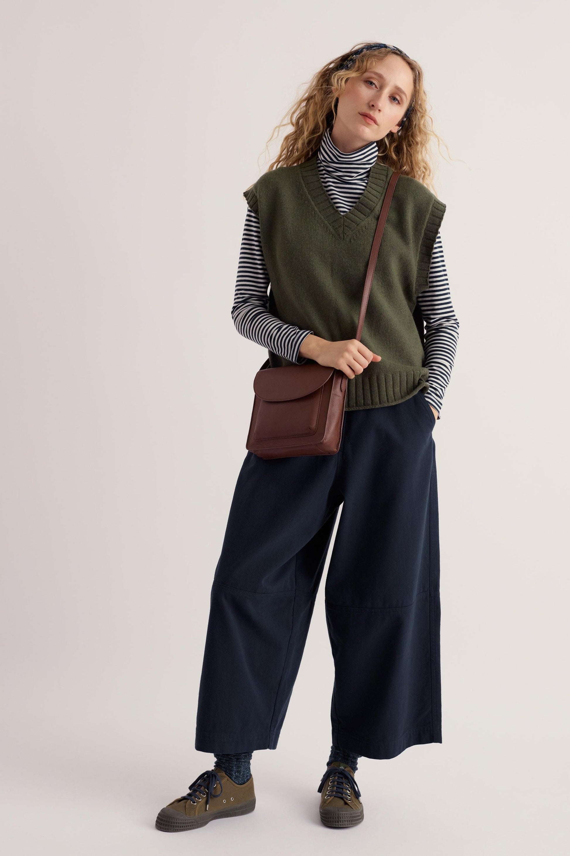 Woman wearing a sustainable khaki knit vest over a striped turtleneck with a brown leather crossbody bag.