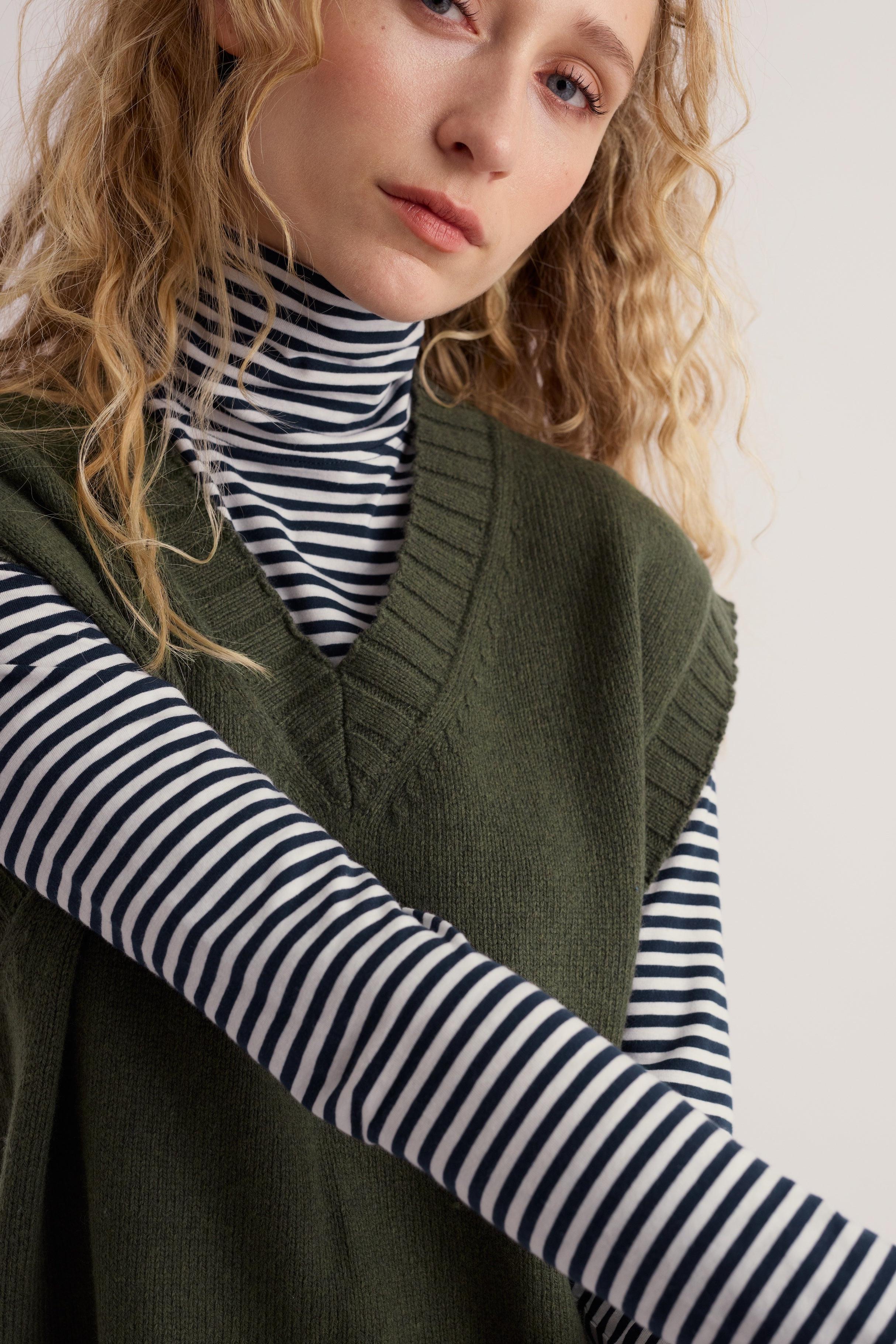 Woman wearing a sustainable khaki knit vest layered over a navy and white striped turtleneck.