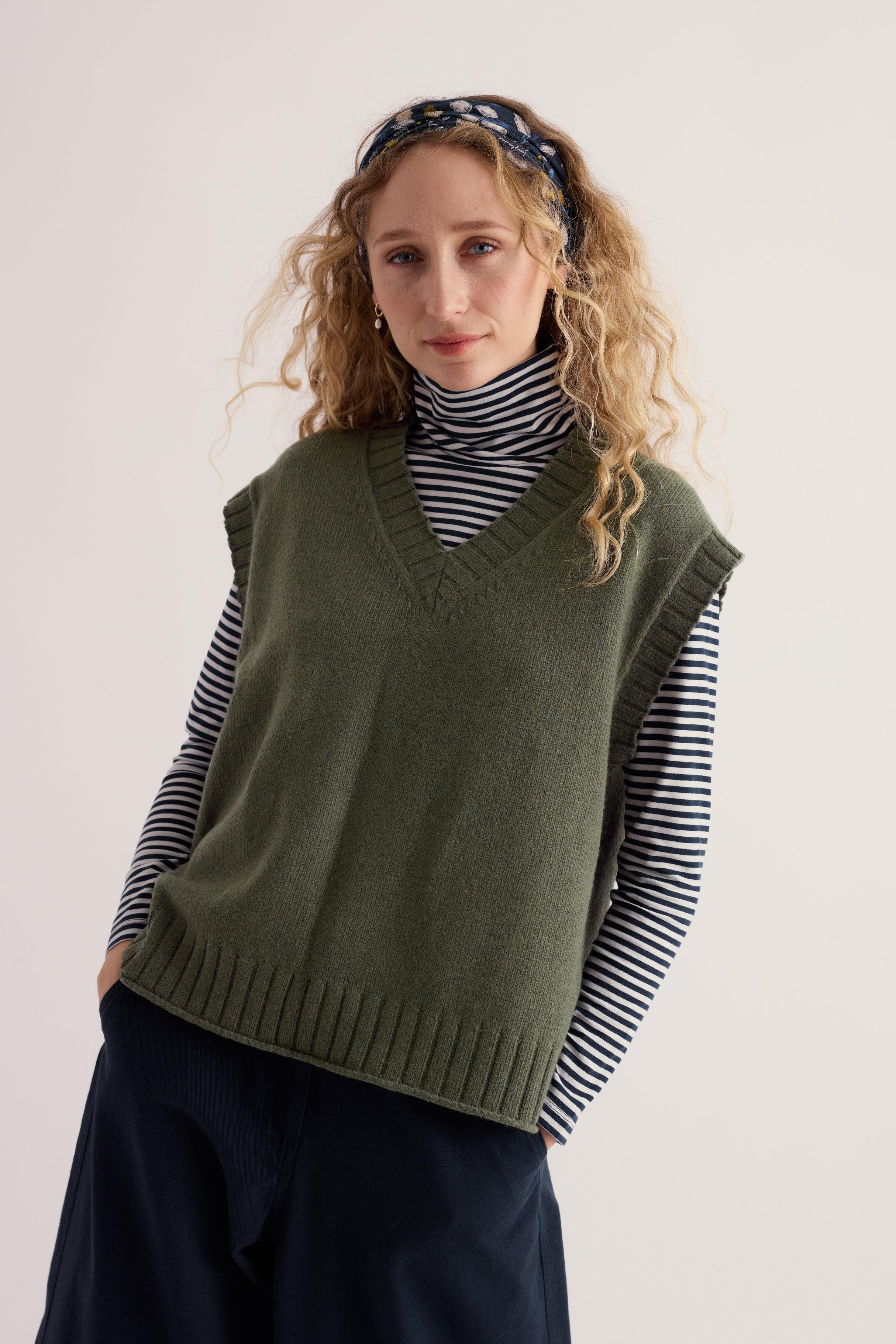 A woman wearing a khaki knit vest over a striped turtleneck with a patterned headband against a plain background.