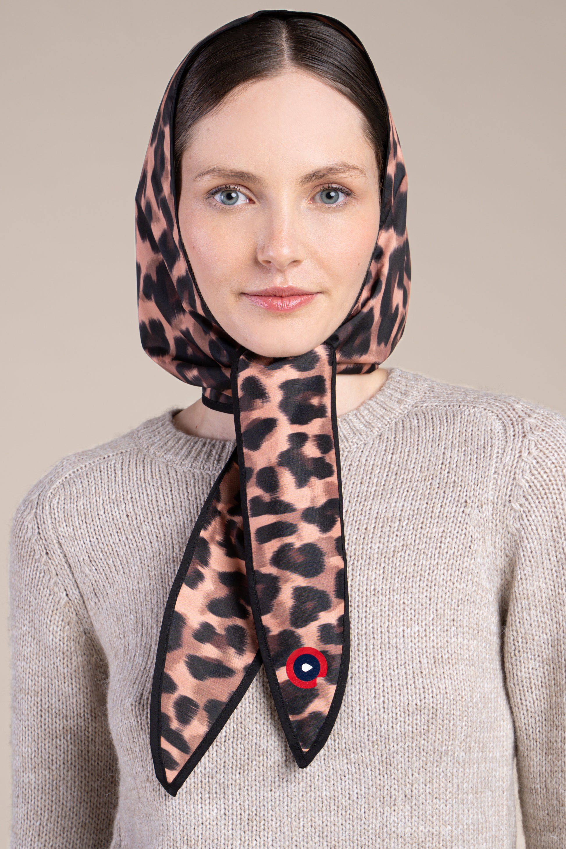 A woman wears a leopard print waterproof head covering tied under the chin against a neutral background.