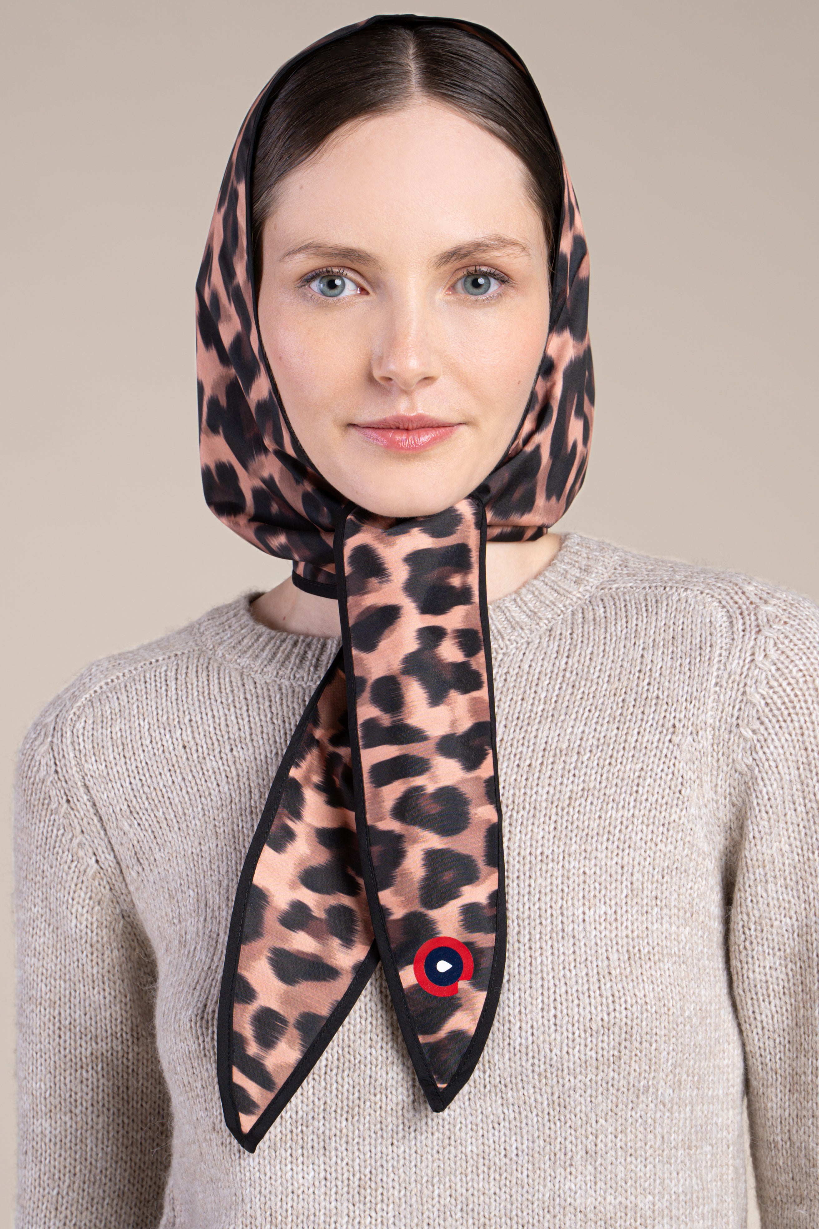 A woman wears a leopard print waterproof head covering tied under the chin against a neutral background.