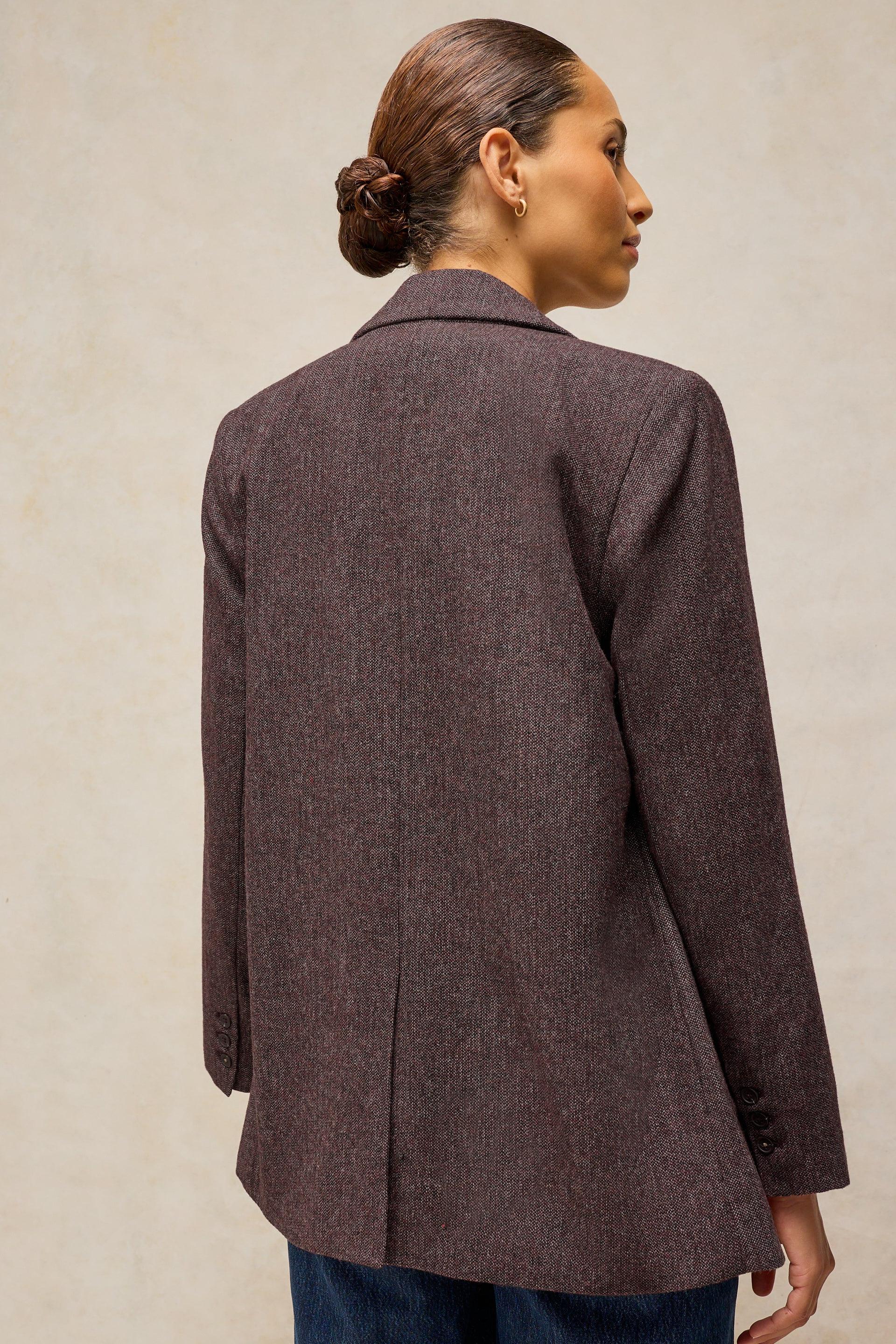 A woman shown from the back wearing a dark brown textured blazer over blue jeans, standing against a neutral beige background.