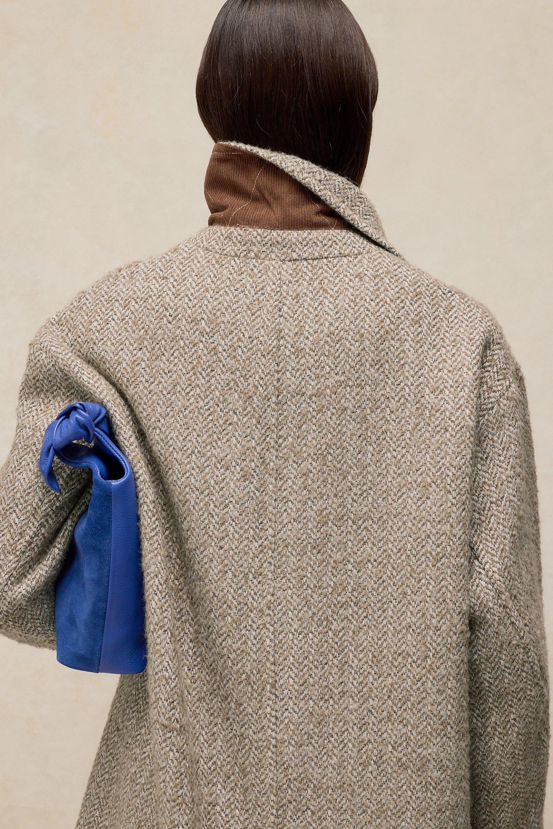 Rear view of a person wearing a textured natural colour wool coat and carrying a bright blue knotted handbag.