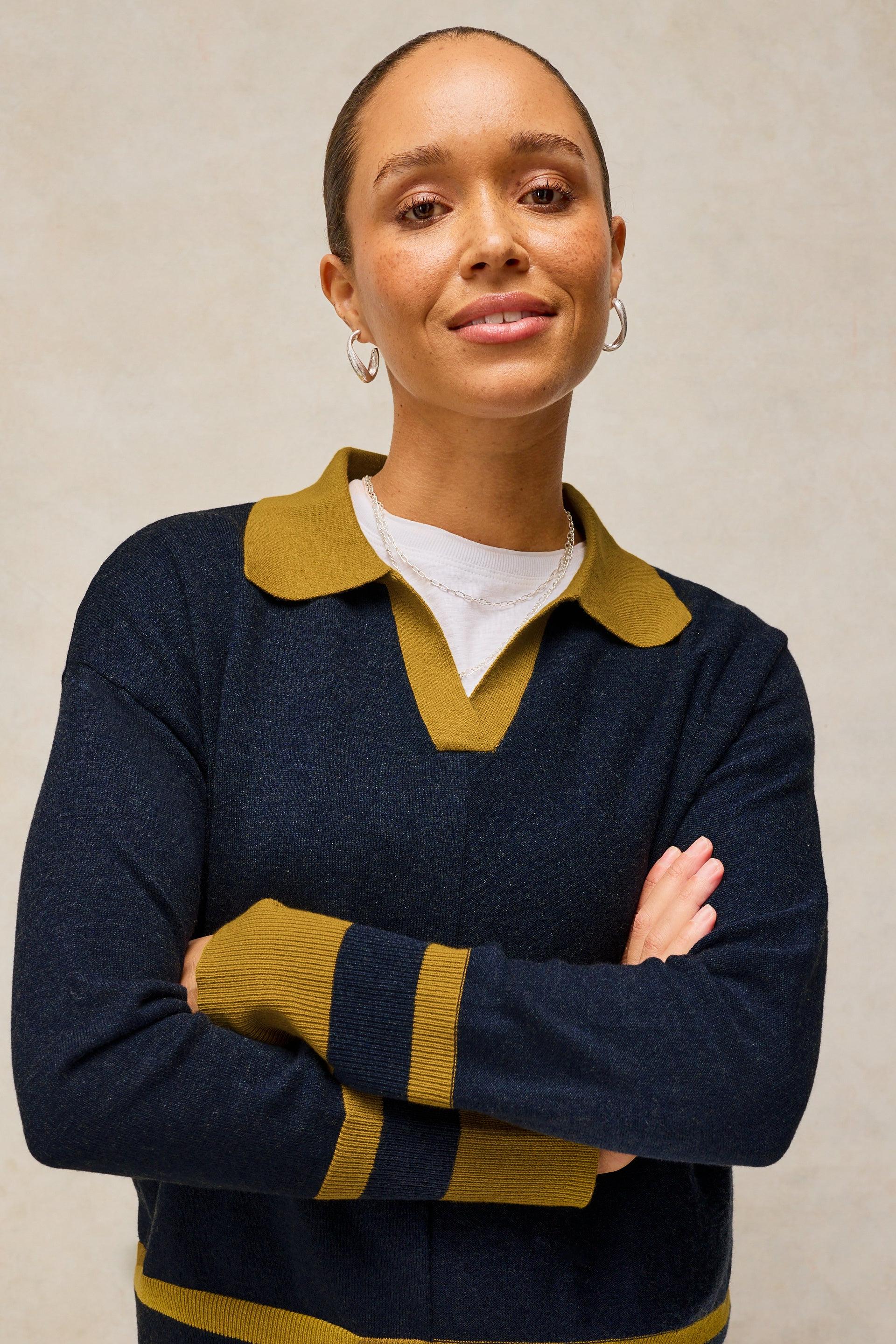 A woman with her hair pulled back wears a navy blue knit sweater with mustard yellow accents on the collar, sleeve cuffs, and hem, layered over a white t-shirt. She is standing with her arms crossed against a neutral textured beige background.