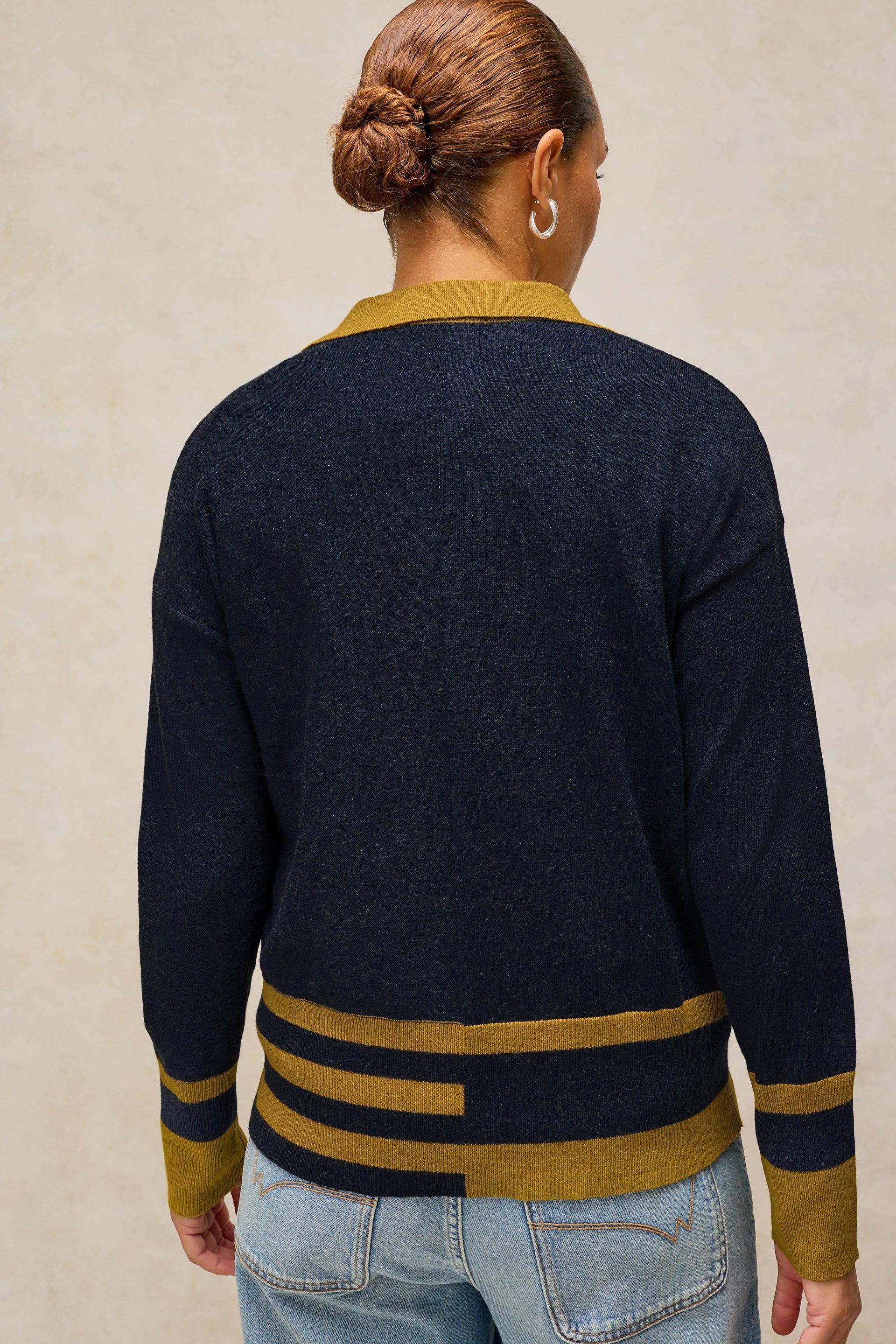 A woman viewed from the back wearing a navy blue knit sweater with mustard yellow stripes and collar, paired with light blue jeans against a neutral beige background.