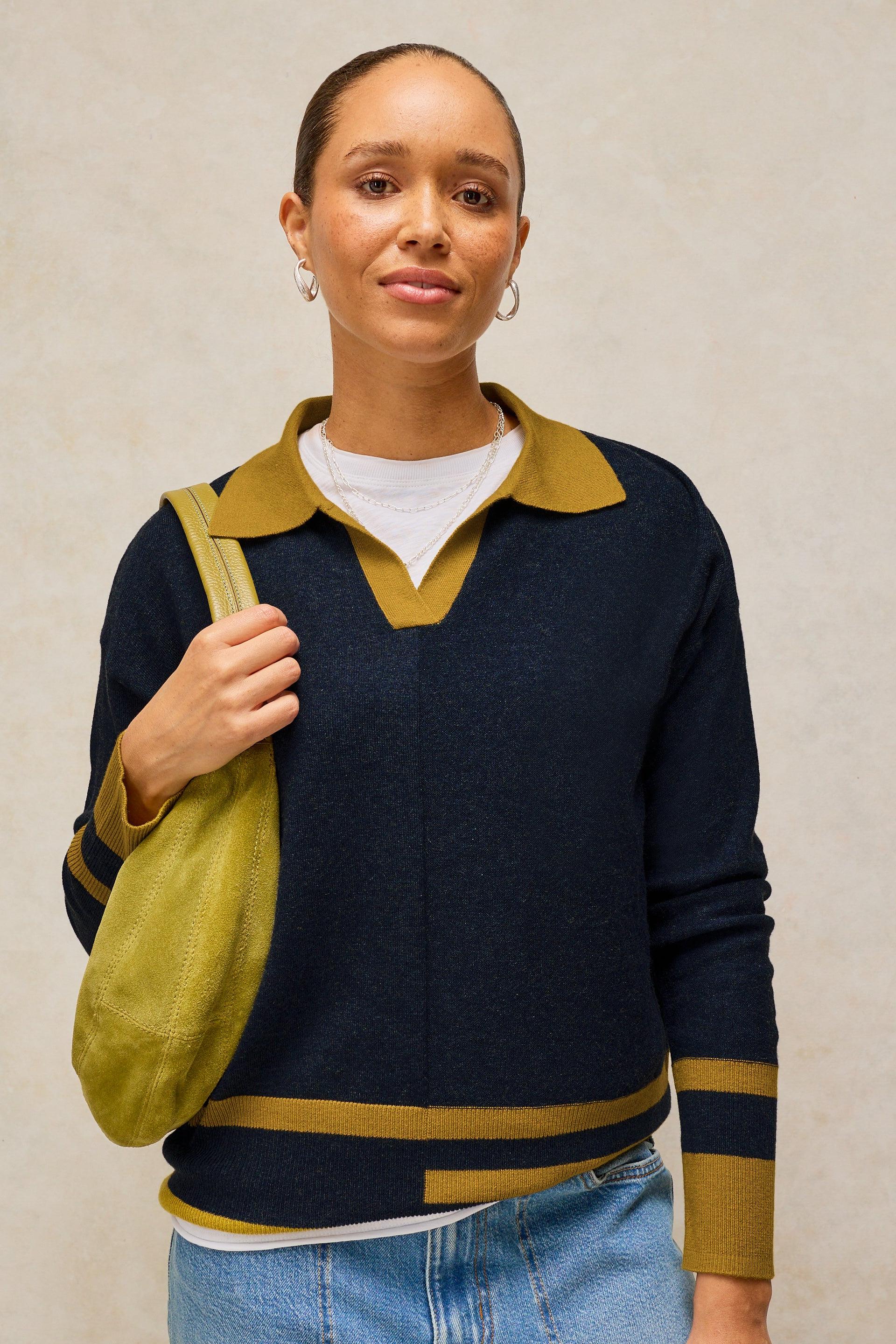 A woman wearing a navy blue knit sweater with mustard yellow collar, cuffs, and hem stripes, layered over a white t-shirt and paired with light blue jeans and a matching yellow shoulder bag, set against a neutral beige background.