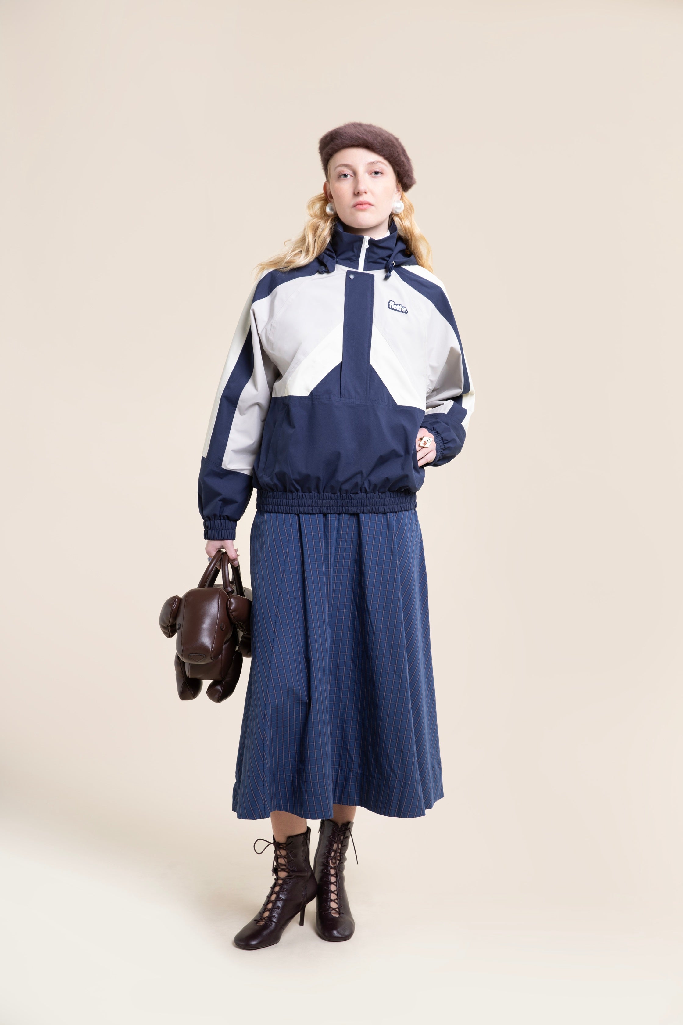 A woman wearing a navy and white waterproof jacket and plaid skirt while holding a brown dog-shaped handbag.