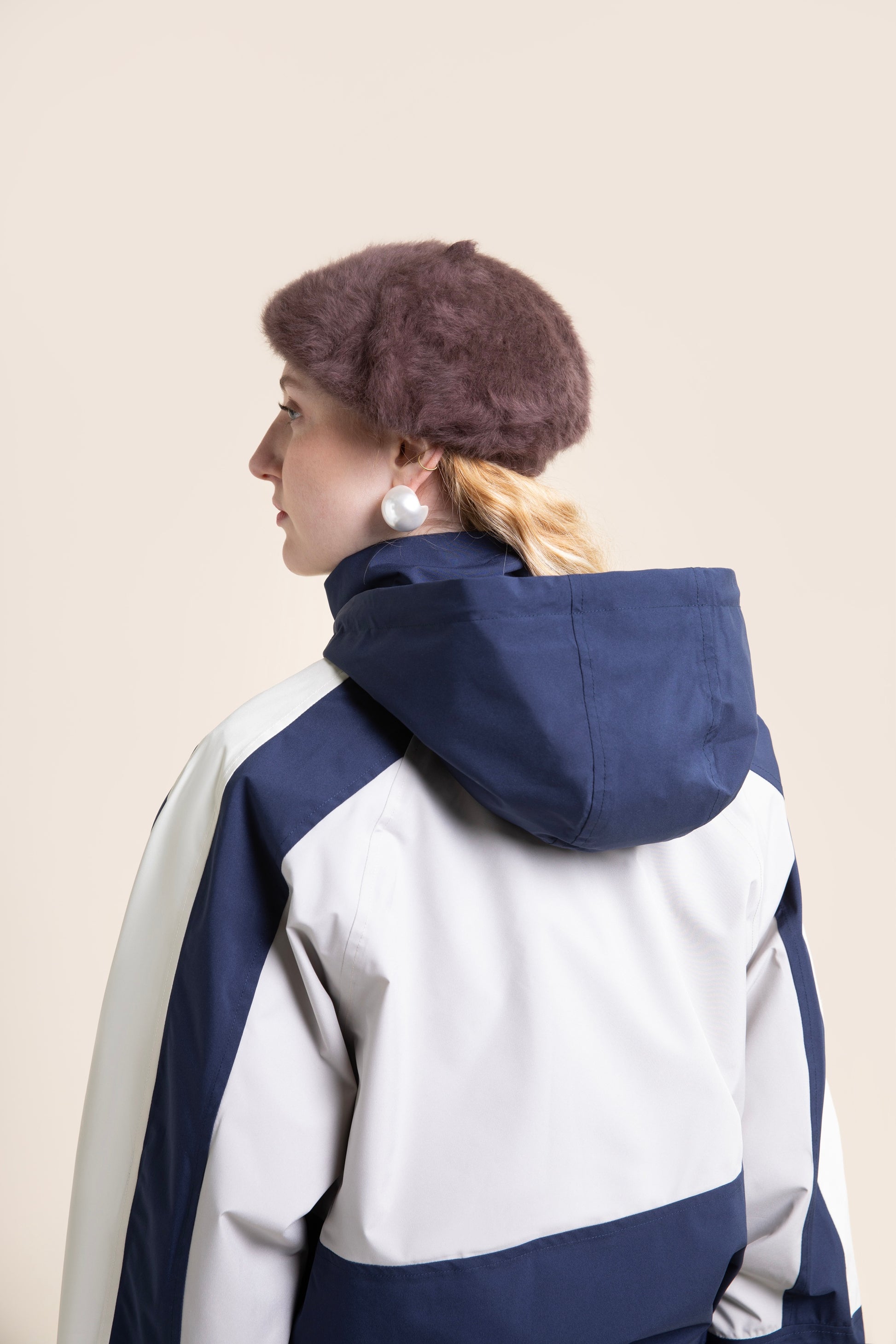 Woman seen from behind wearing a navy and white waterproof jacket and a brown beret against a neutral background.