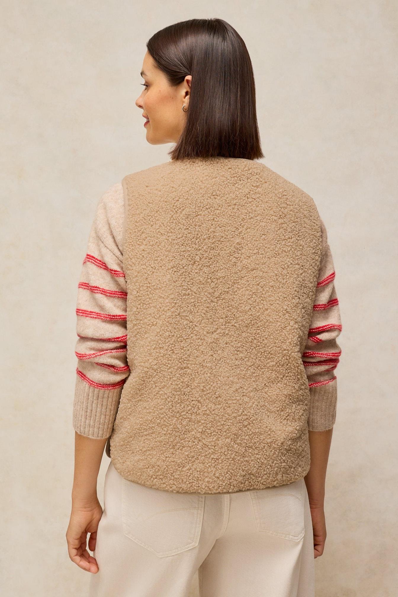 A woman wearing a cosy neutral colour fleece gilet over a striped sweater against a plain background.