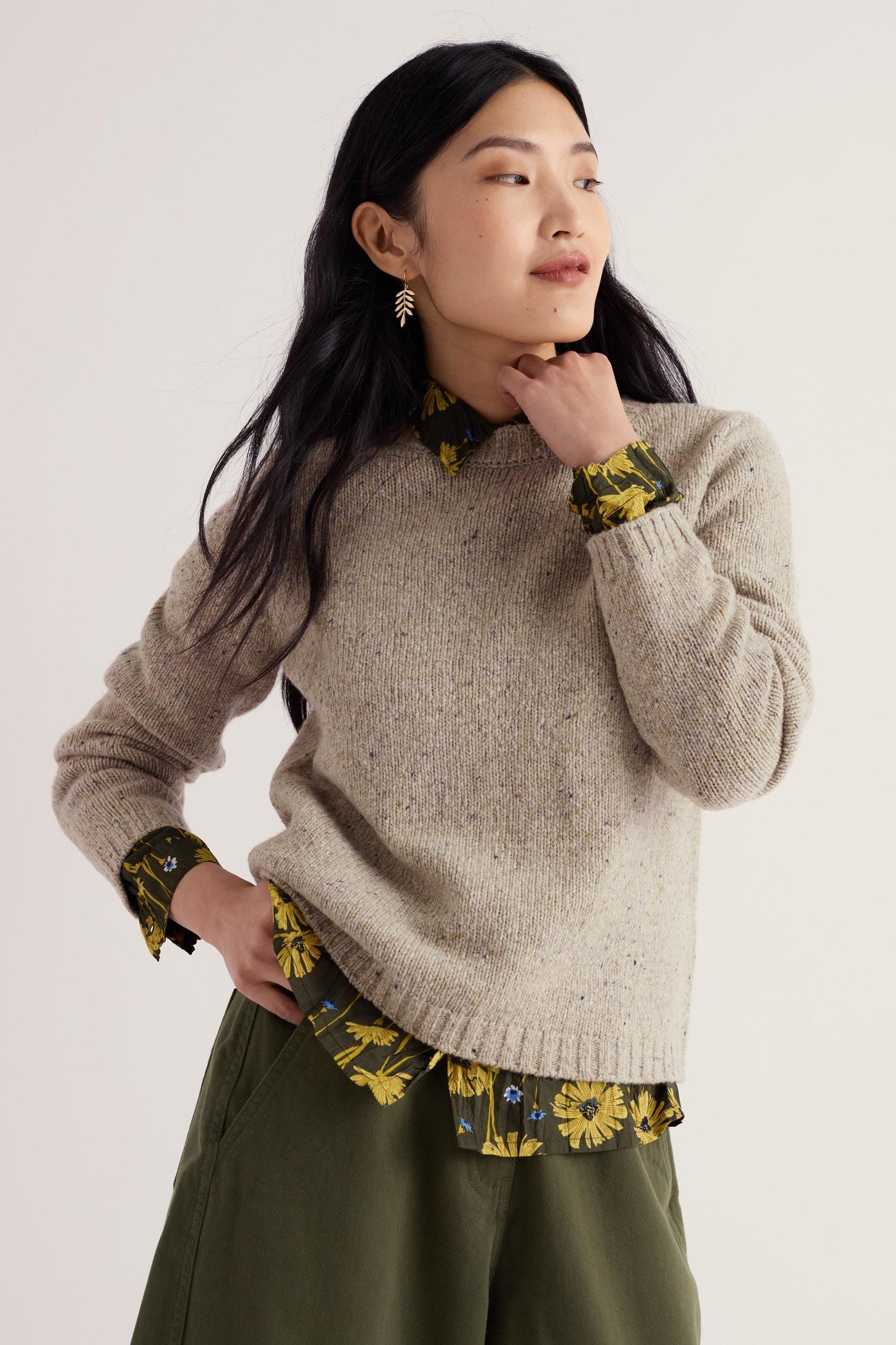 A woman wearing a neutral merino wool jumper layered over a dark floral shirt against a plain background.