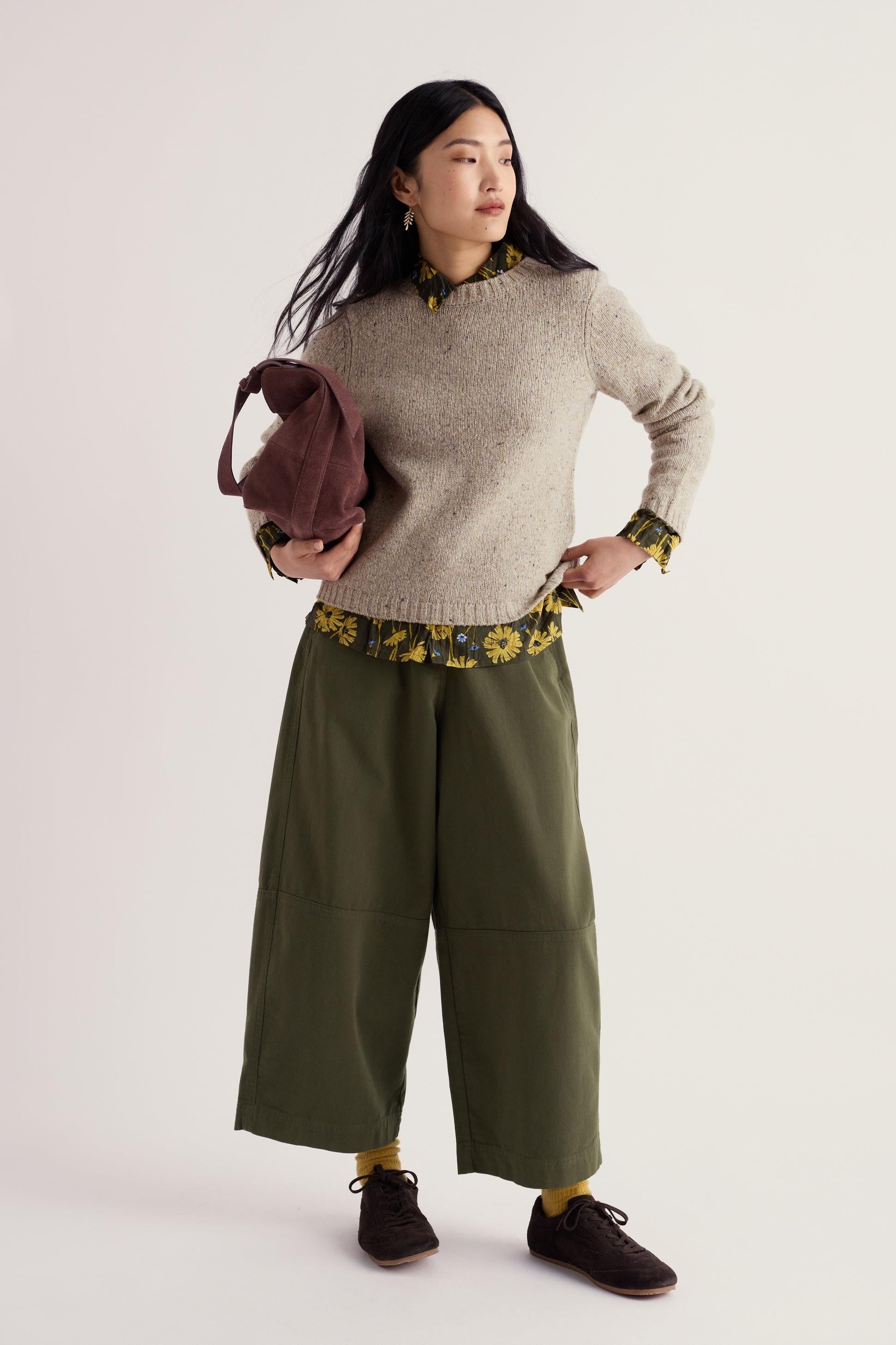 A woman posing in a neutral wool jumper over a floral shirt and wide-leg olive trousers.