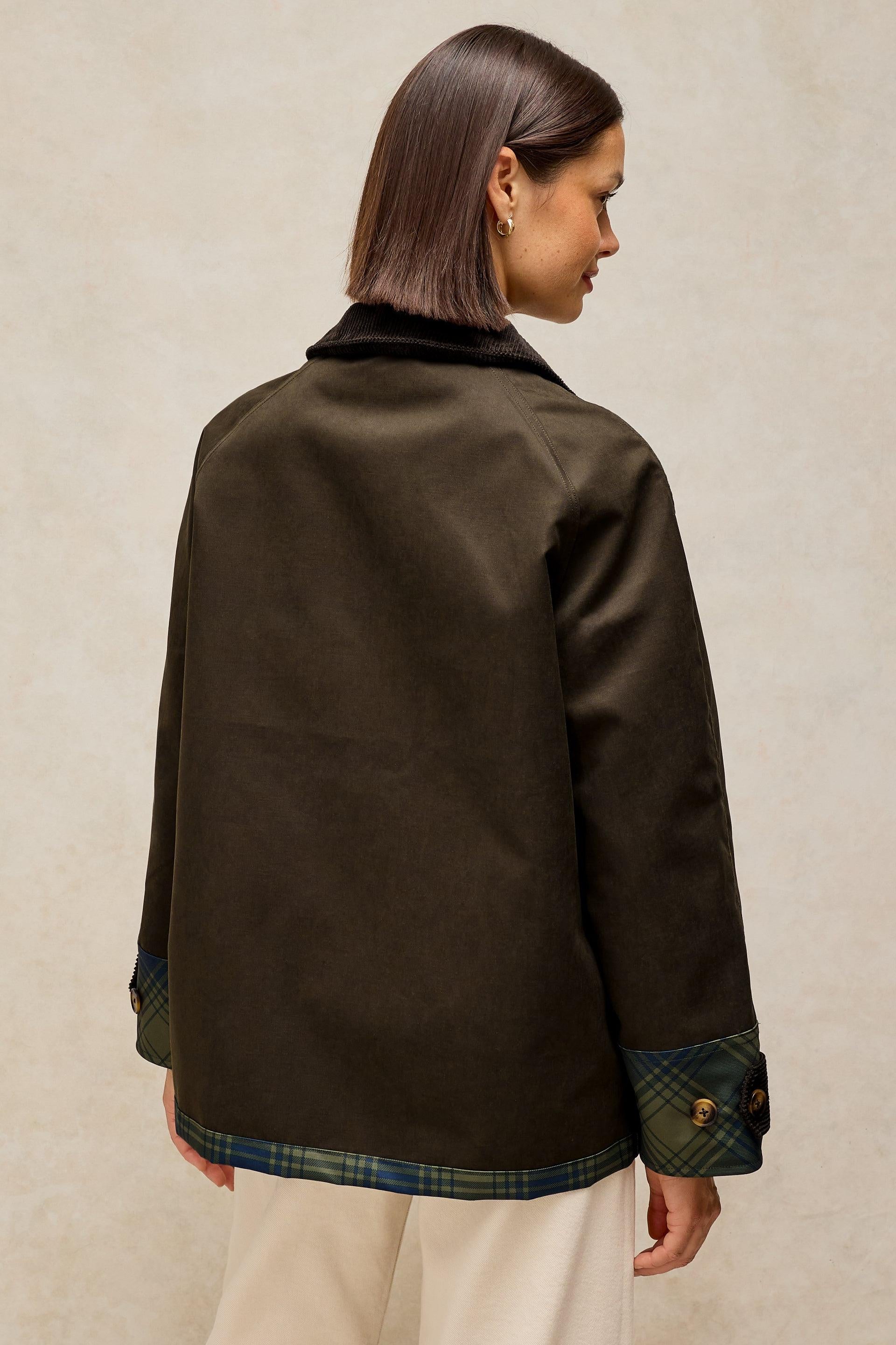 A woman shown from the back wearing a dark brown jacket with a green plaid lining at the cuffs and hem, paired with cream trousers, against a textured beige background.