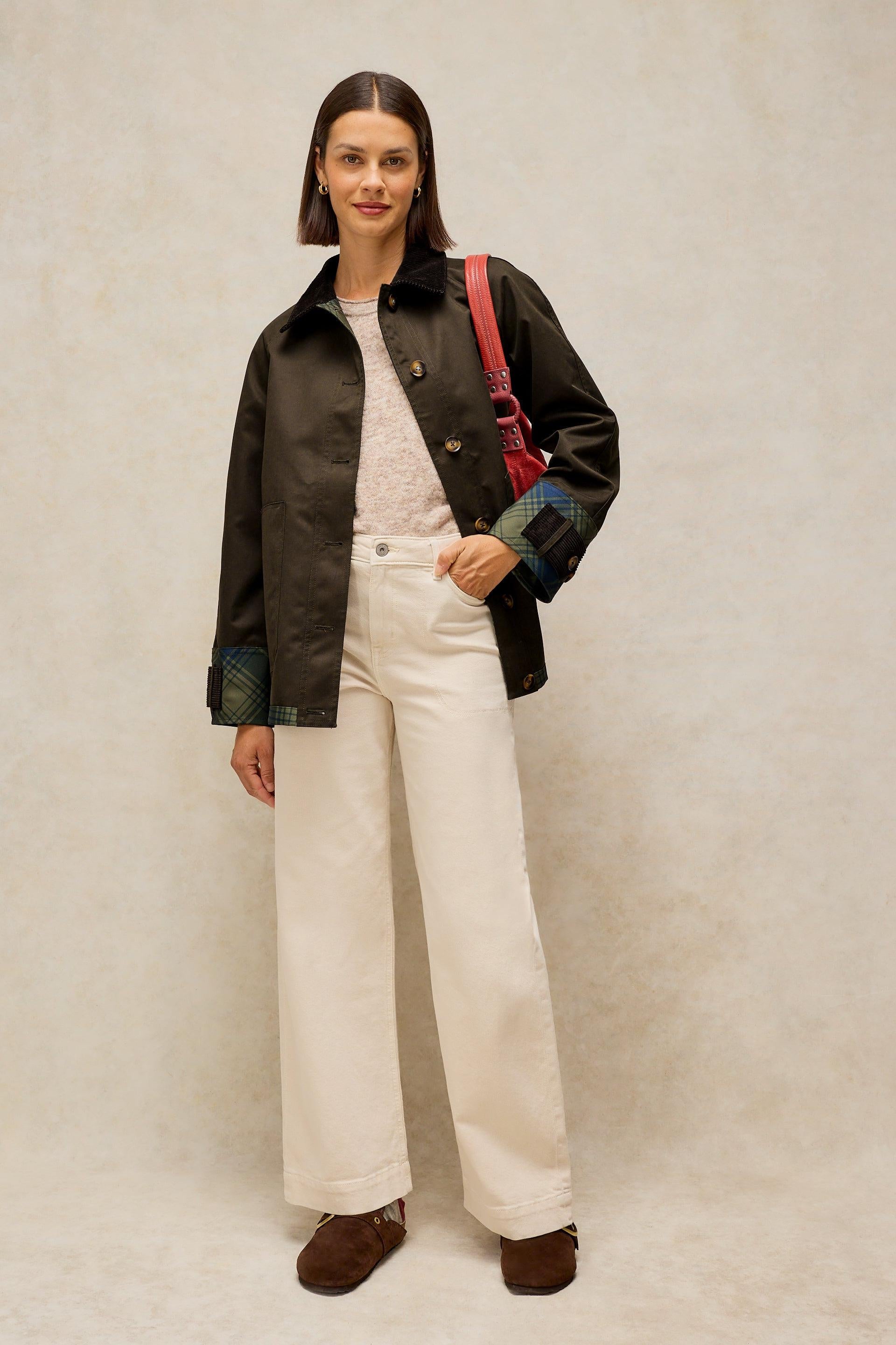 A woman stands against a textured beige studio backdrop wearing a black waxed jacket with green plaid cuffs, off-white wide-leg trousers, brown shoes, and a red shoulder bag.
