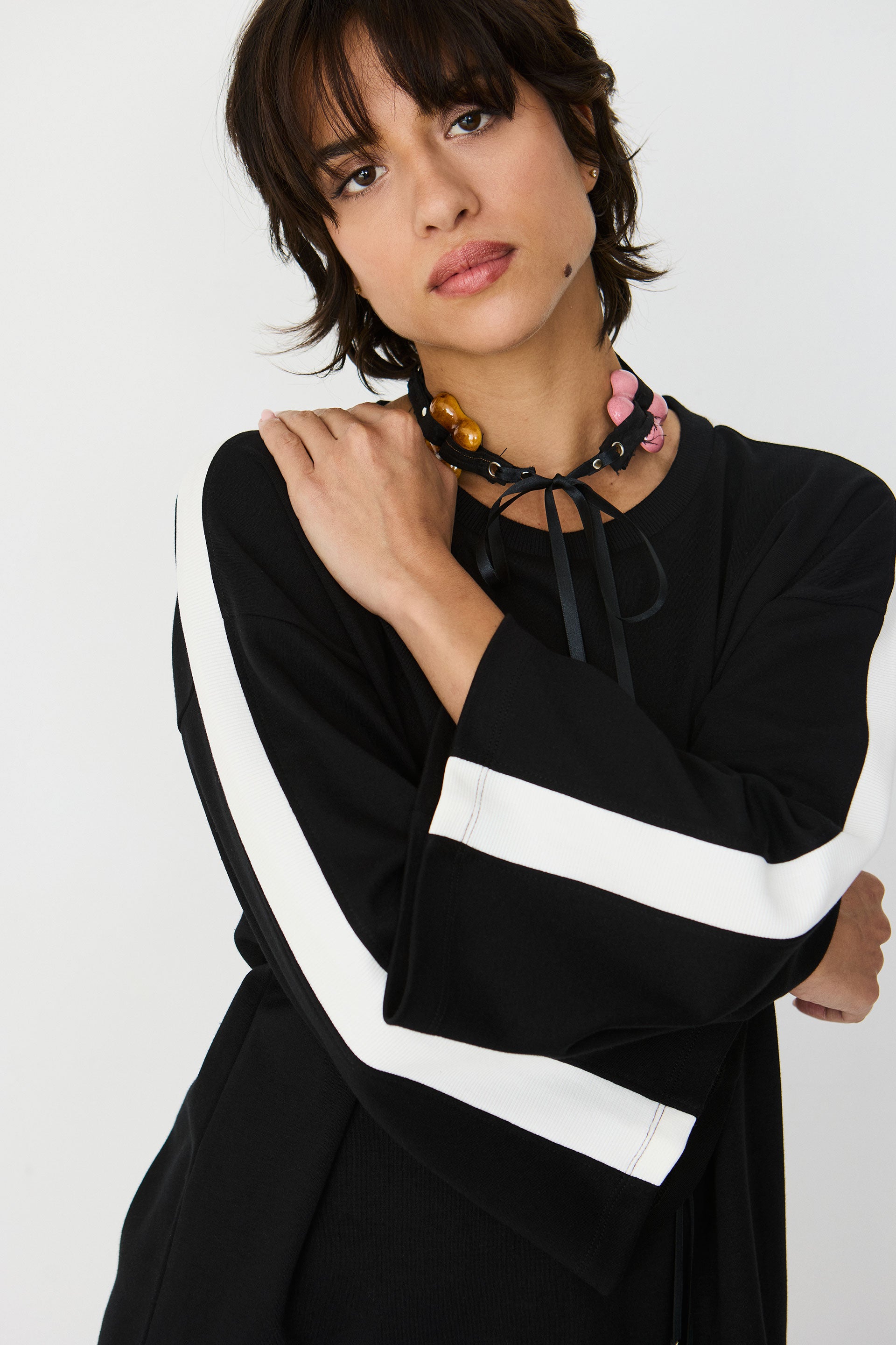 A woman wearing a black long-sleeve top with bold white stripes on the sleeves, styled with a chunky beaded necklace, against a plain light gray background.
