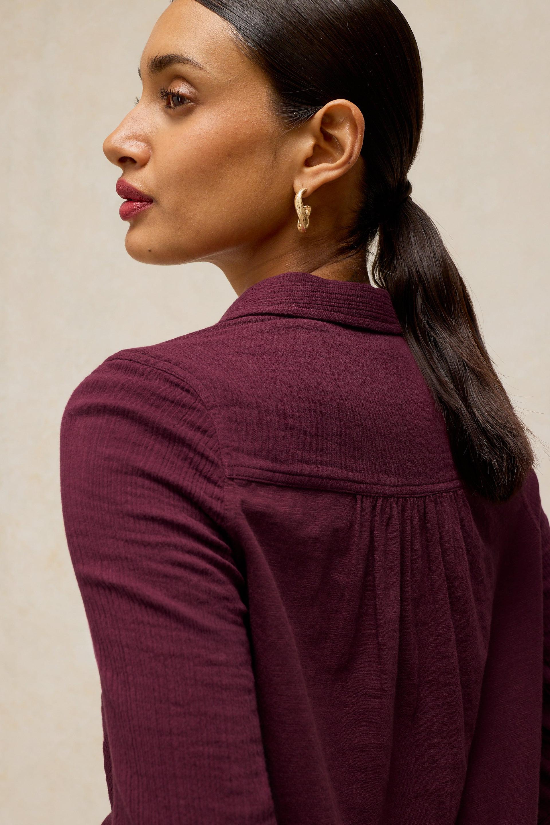 A woman shown from the back and side wearing a textured deep plum-purple long-sleeve blouse against a neutral beige background.