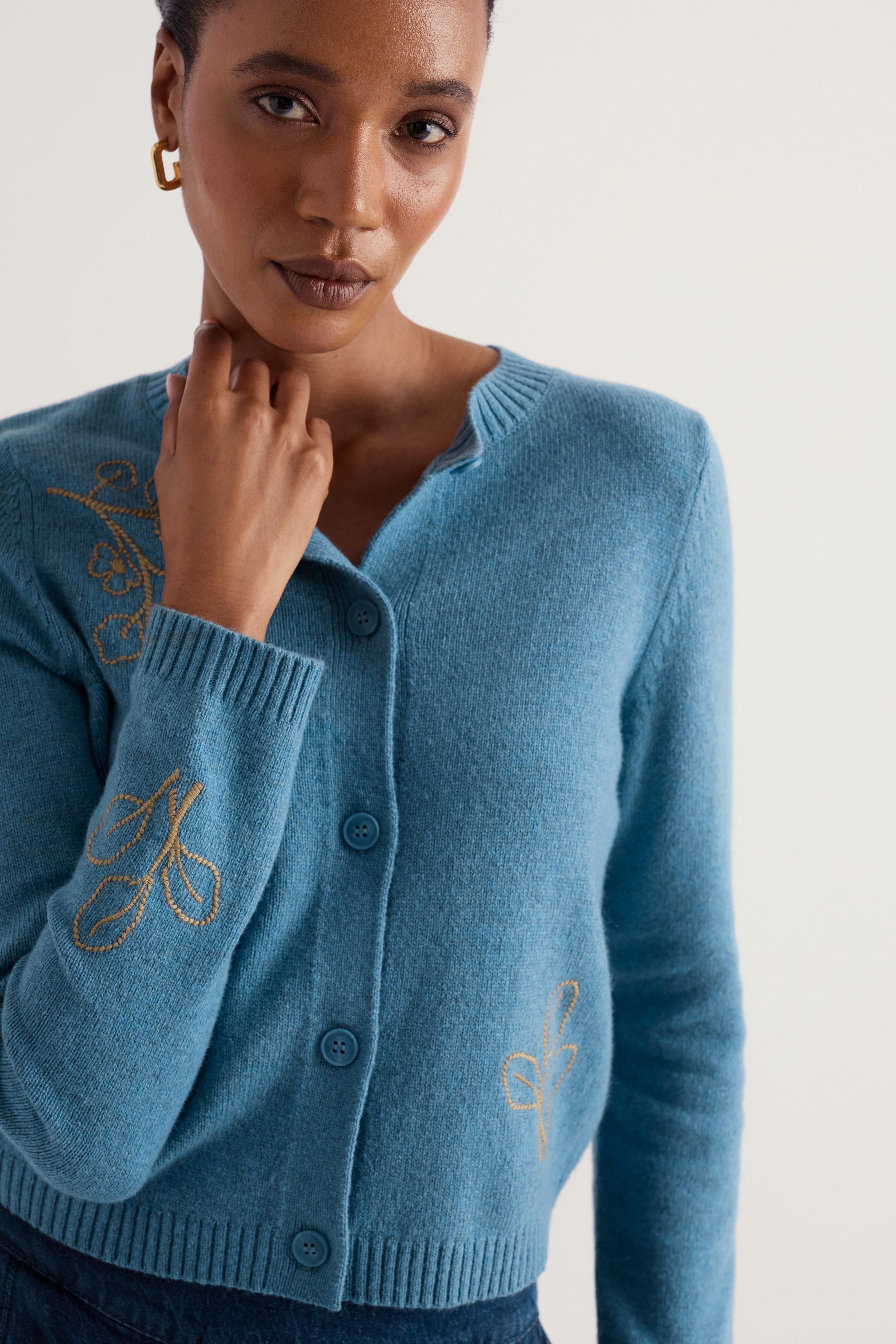A woman wearing a light blue knit cardigan sweater with gold embroidered script details on the sleeves and chest, posed against a plain off-white background.