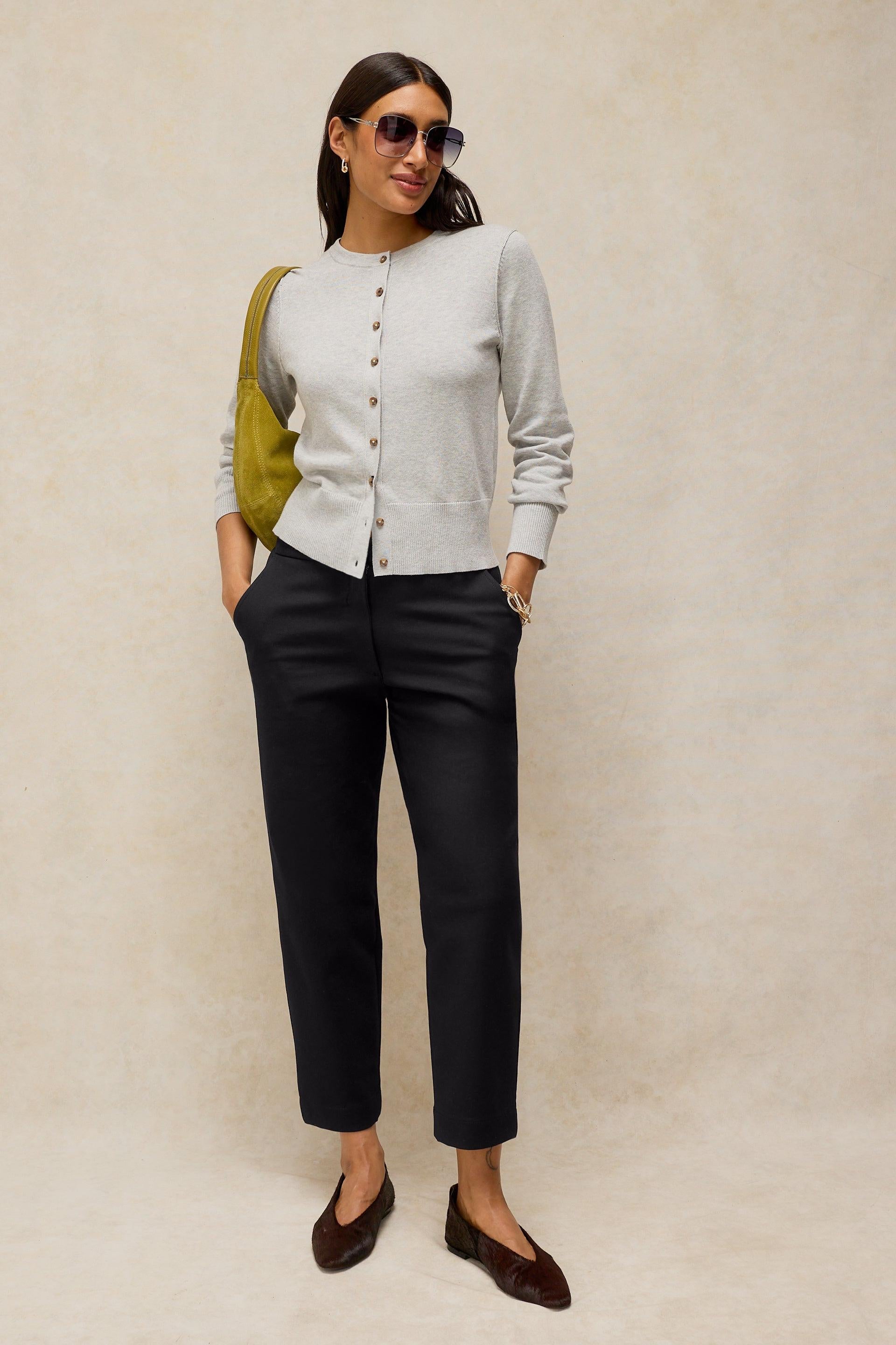 A woman modeling a light gray long-sleeve button-front cardigan, black cropped trousers, and dark brown flats, carrying a yellow-green shoulder bag against a textured beige studio backdrop.