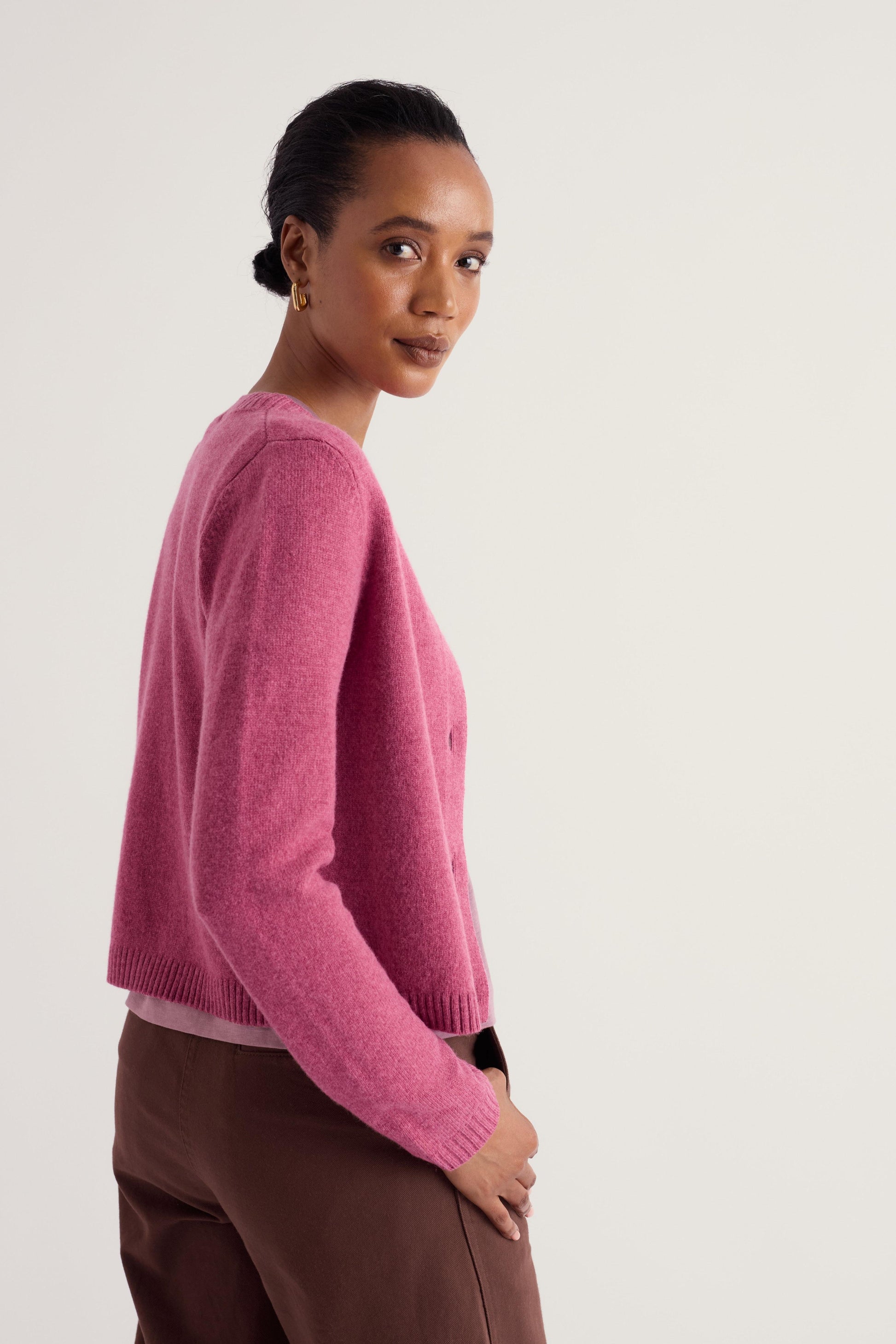 A woman wearing a pink knit cardigan and brown trousers, photographed from the side against a plain light gray studio background.