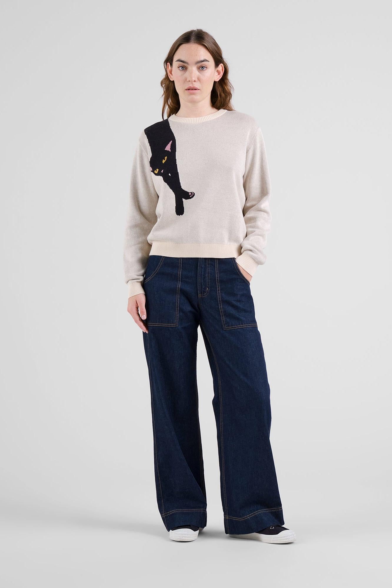 A model stands against a light gray studio background wearing a cream-colored crewneck sweater with a black cat graphic on the chest, paired with dark blue wide-leg jeans and white sneakers.