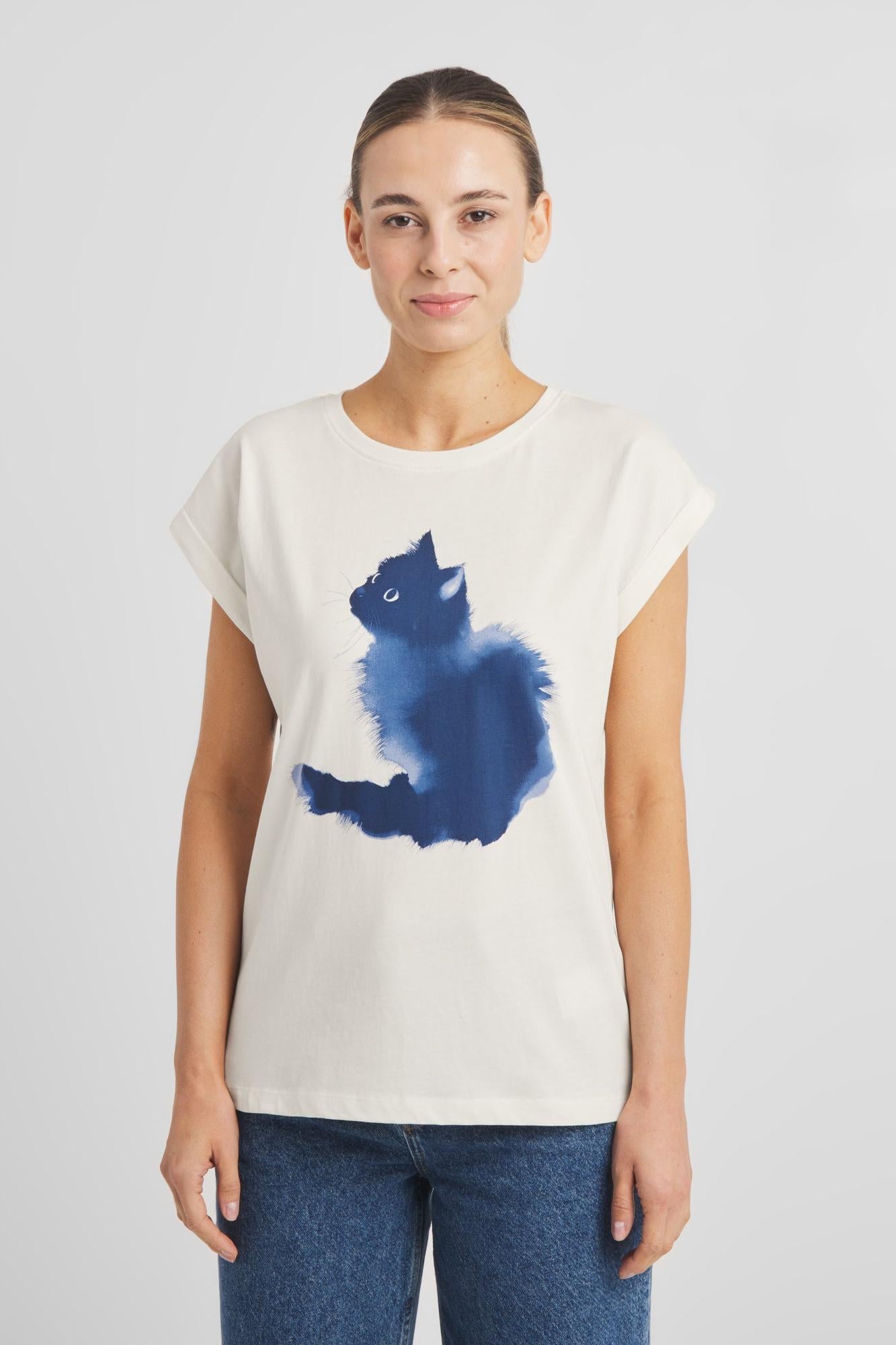 A woman modeling a cream-colored short-sleeve t-shirt with a blue watercolor-style cat graphic on the front, paired with blue jeans, against a light gray studio background.
