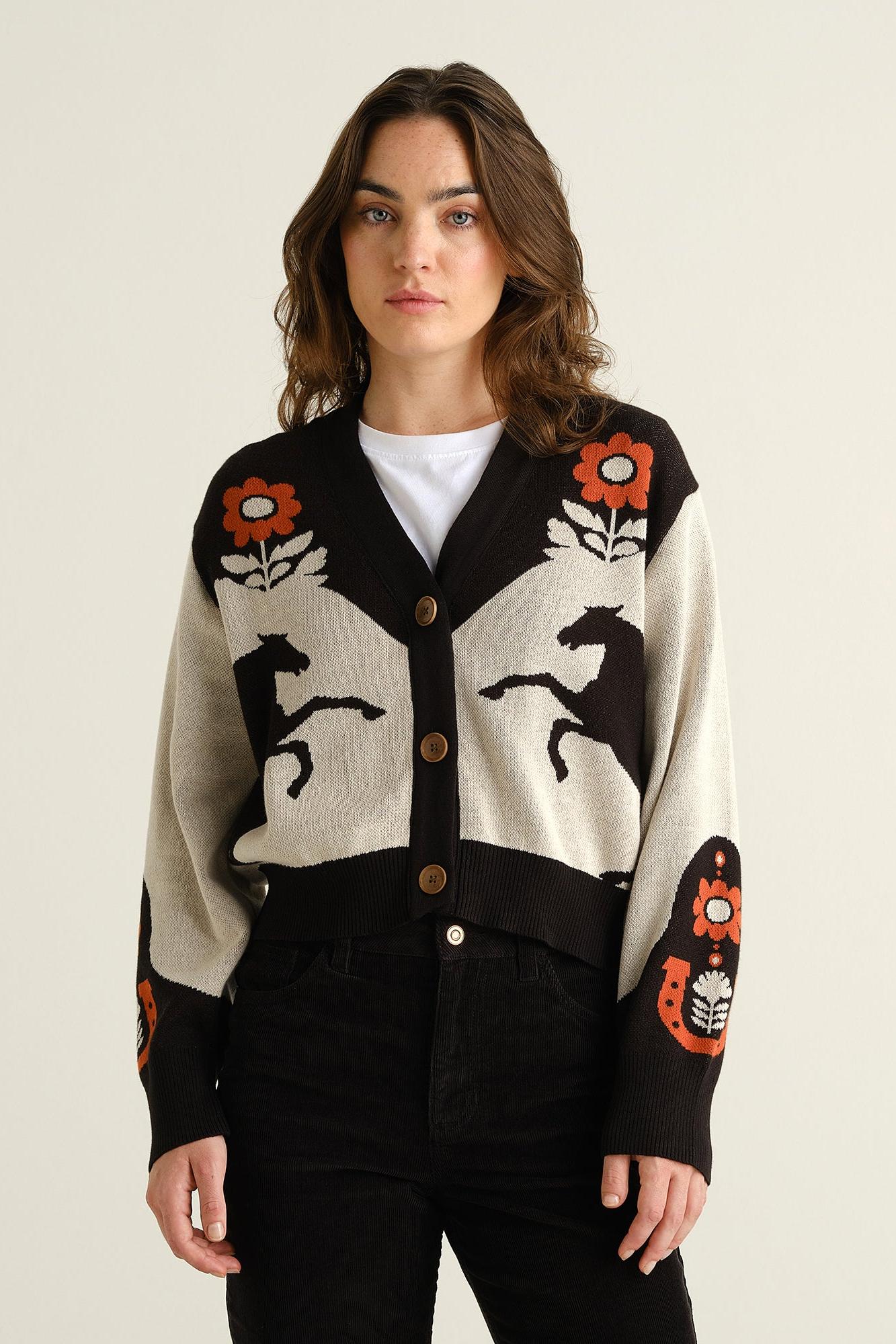 A woman modeling a cropped cardigan sweater with a cream base, black trim and motifs, and orange floral accents, paired with a white t-shirt and black jeans against a light beige background.