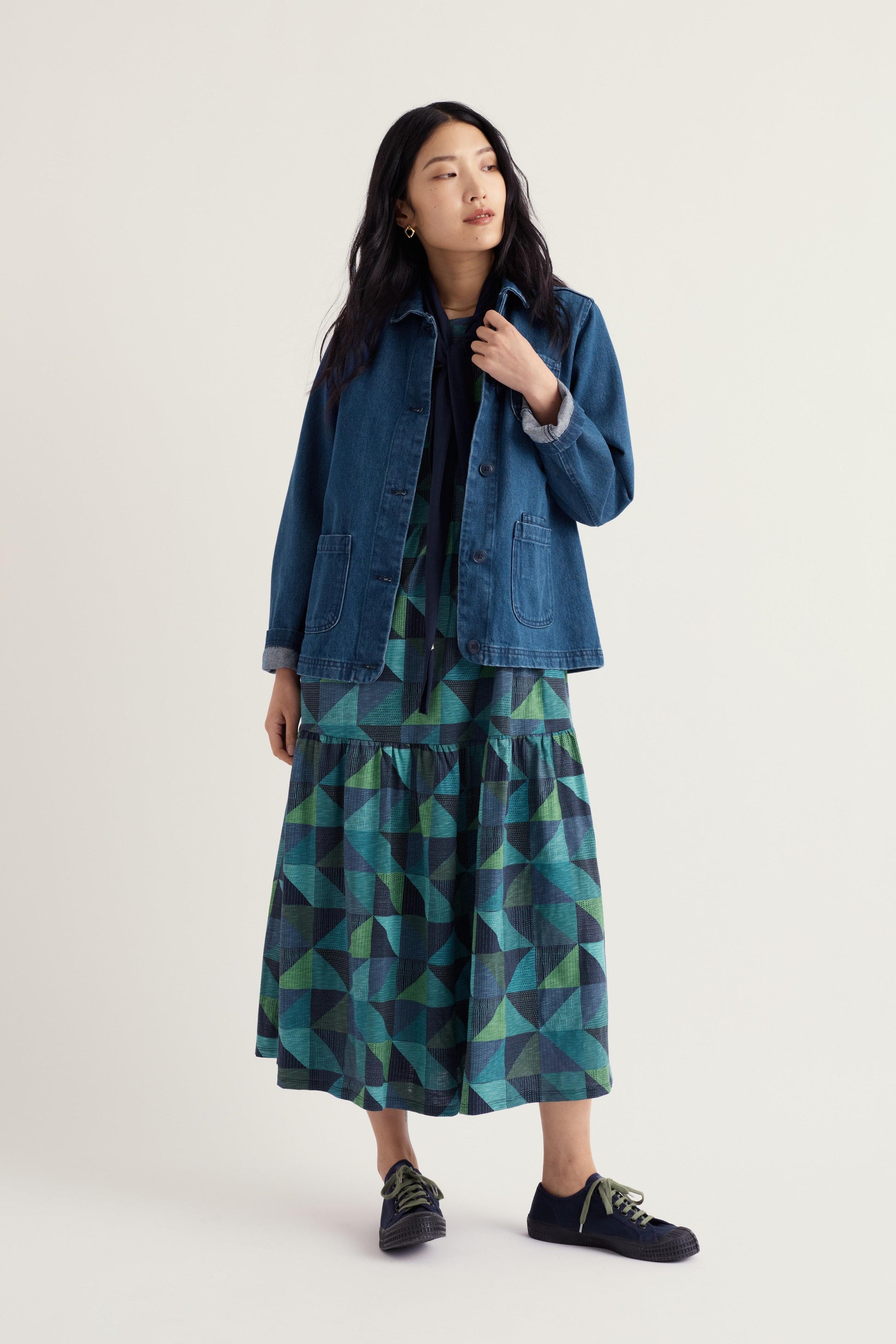 A woman modeling a blue denim jacket over a black top and a tiered midi skirt with a green and blue geometric triangle pattern, paired with black slip-on shoes, against a light gray studio background.