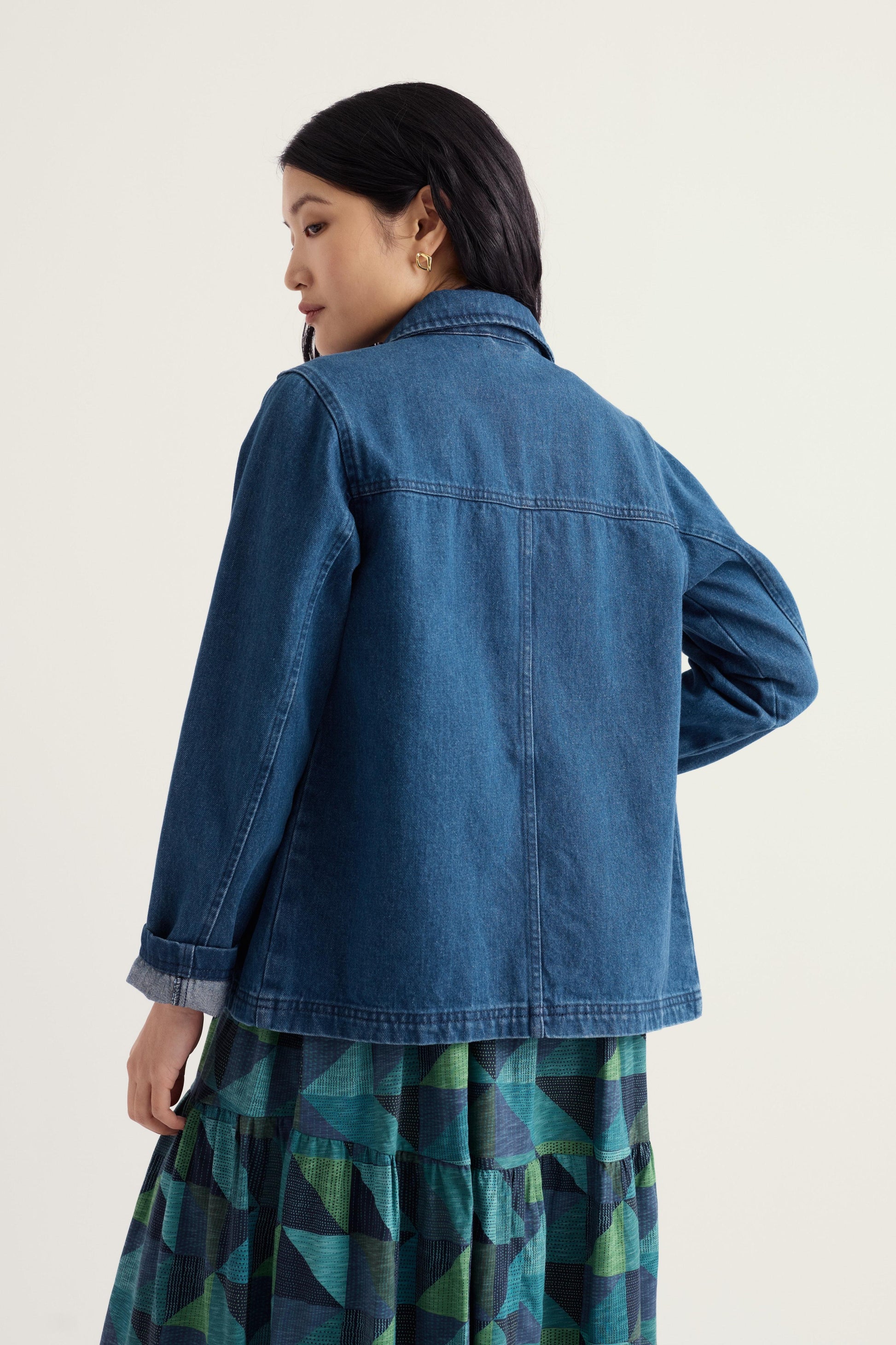 A woman shown from the back wearing a blue denim jacket over a green and blue geometric patterned skirt, photographed against a plain off-white studio background.