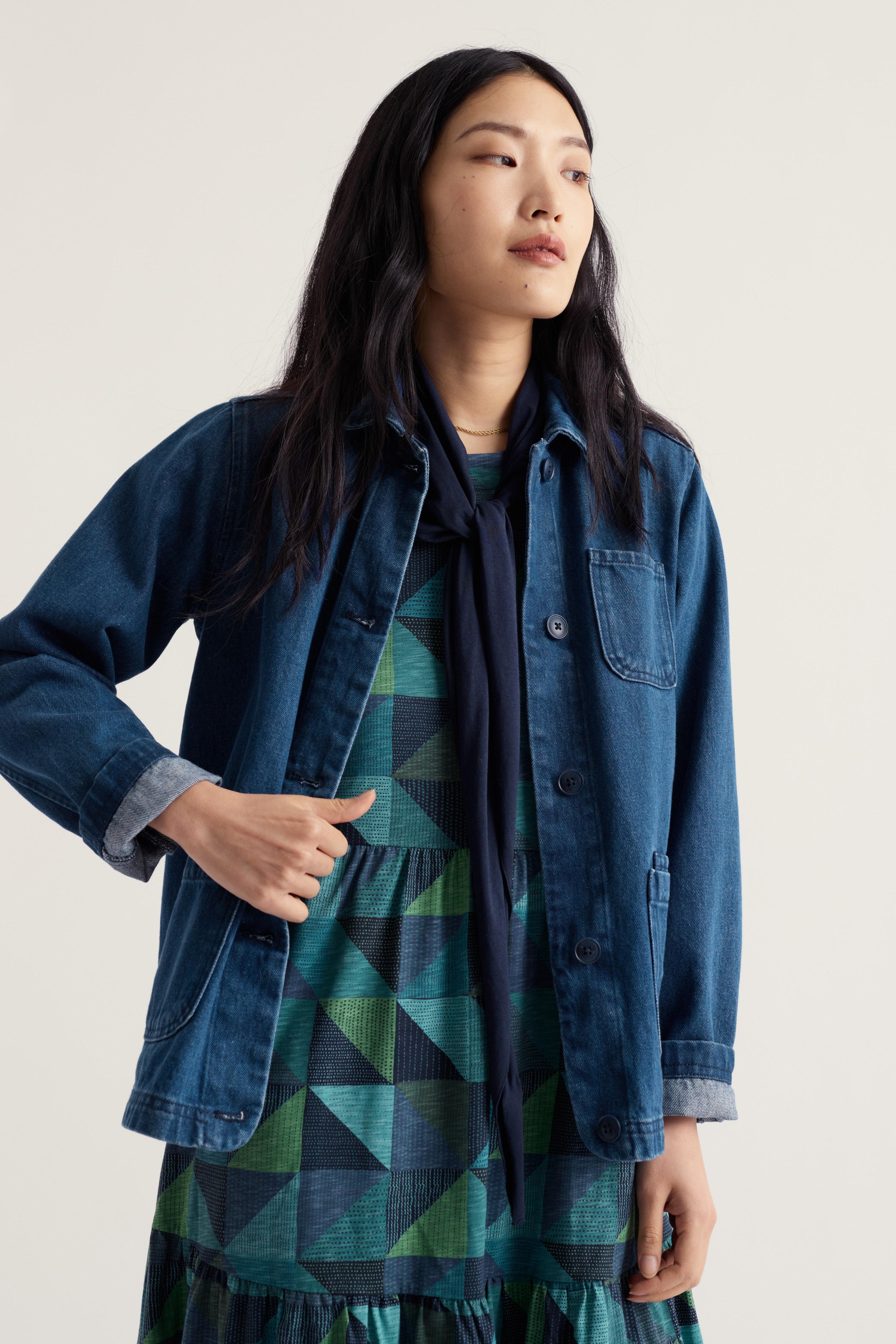 A woman wearing a dark blue denim button-up shirt jacket over a green and navy geometric patterned dress, photographed against a plain light beige studio background.