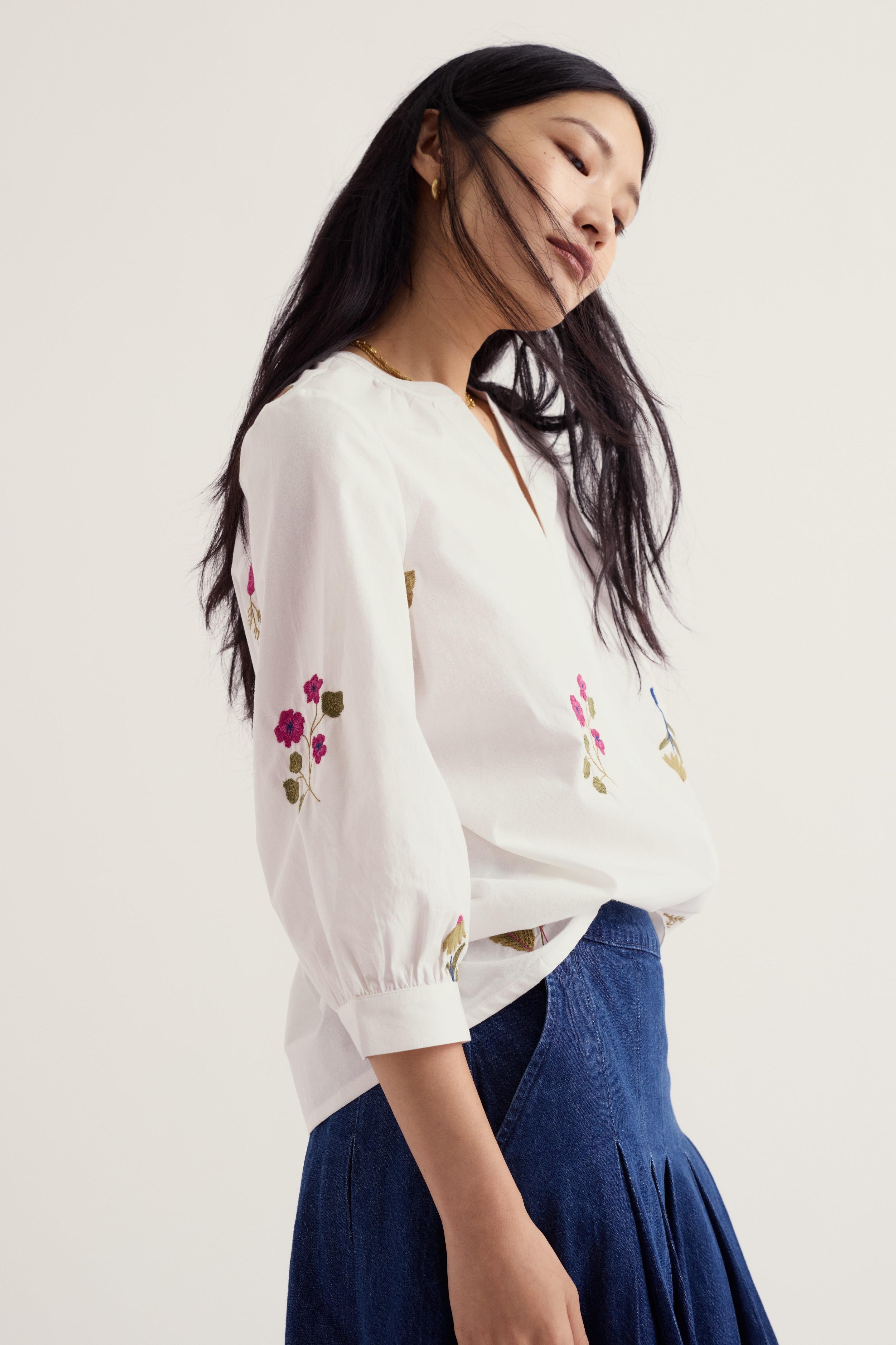 A woman wearing a white long-sleeve blouse with colorful floral embroidery on the sleeves and a dark blue skirt, photographed against a plain light background.