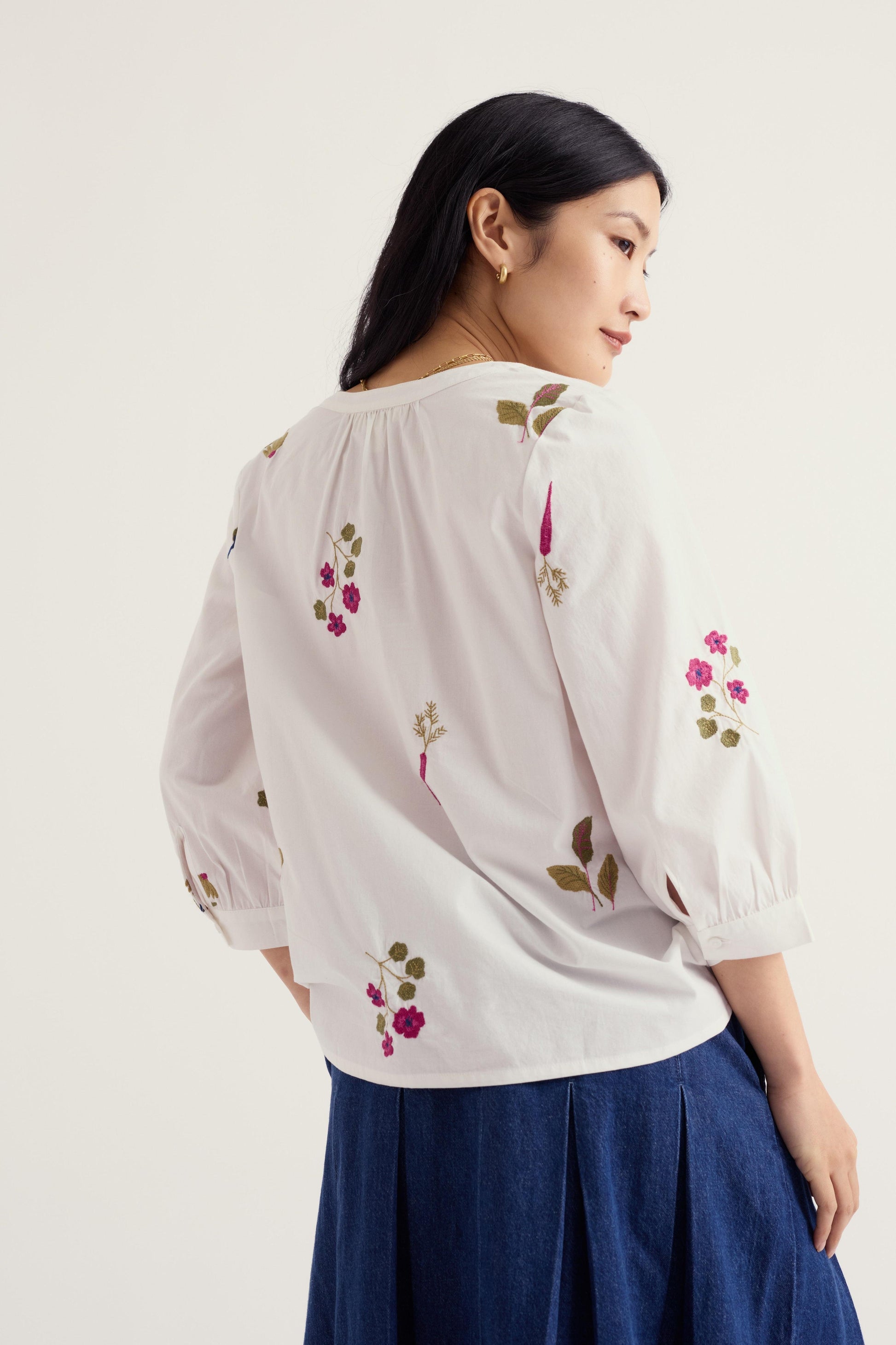 A woman shown from the back wearing a white blouse with pink and green floral embroidery and a dark blue skirt, photographed against a plain light background.