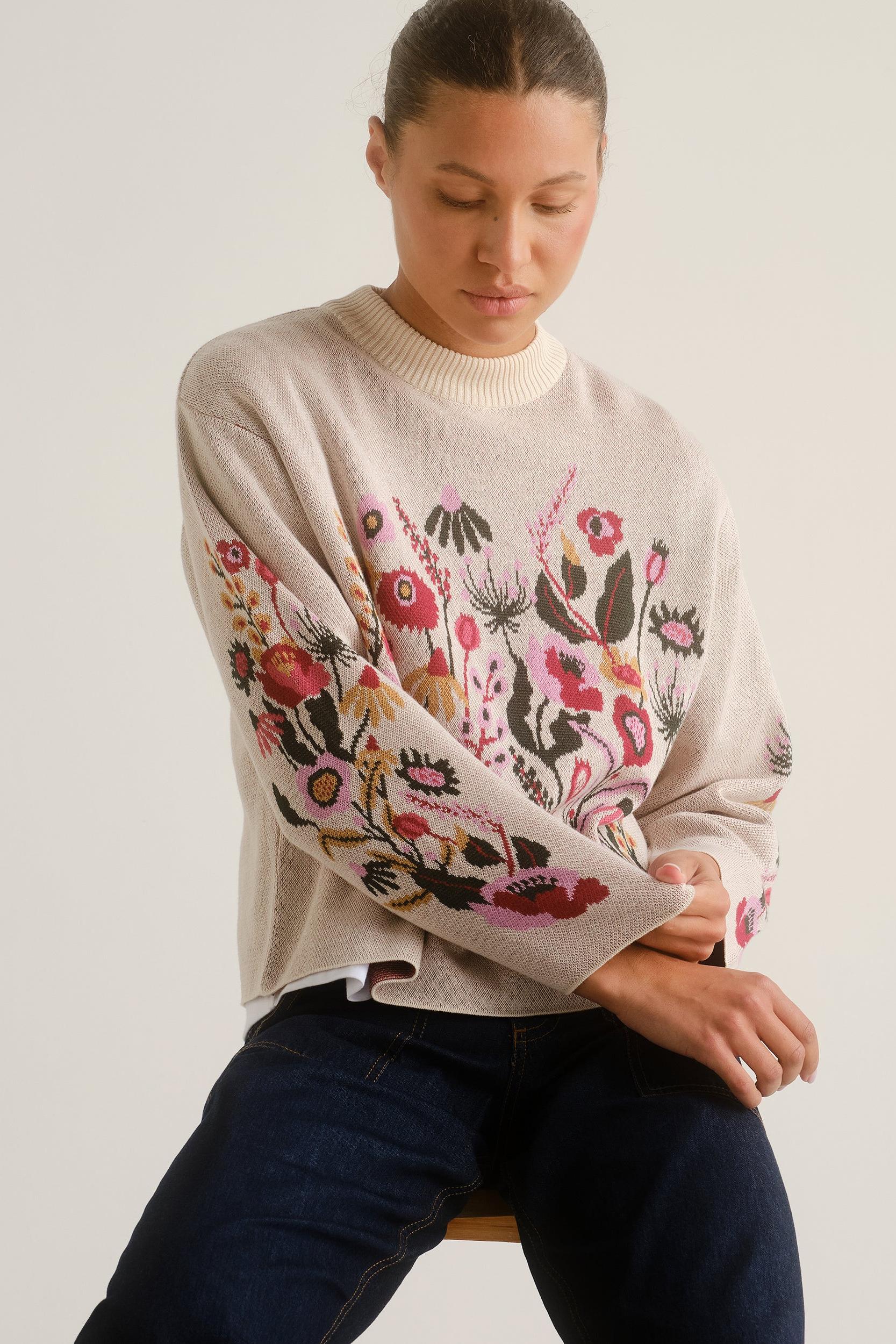 A woman wearing a cream-colored knit sweater with a vibrant, multicolored floral and botanical pattern across the front and sleeves, paired with dark blue jeans, seated against a plain light beige background.